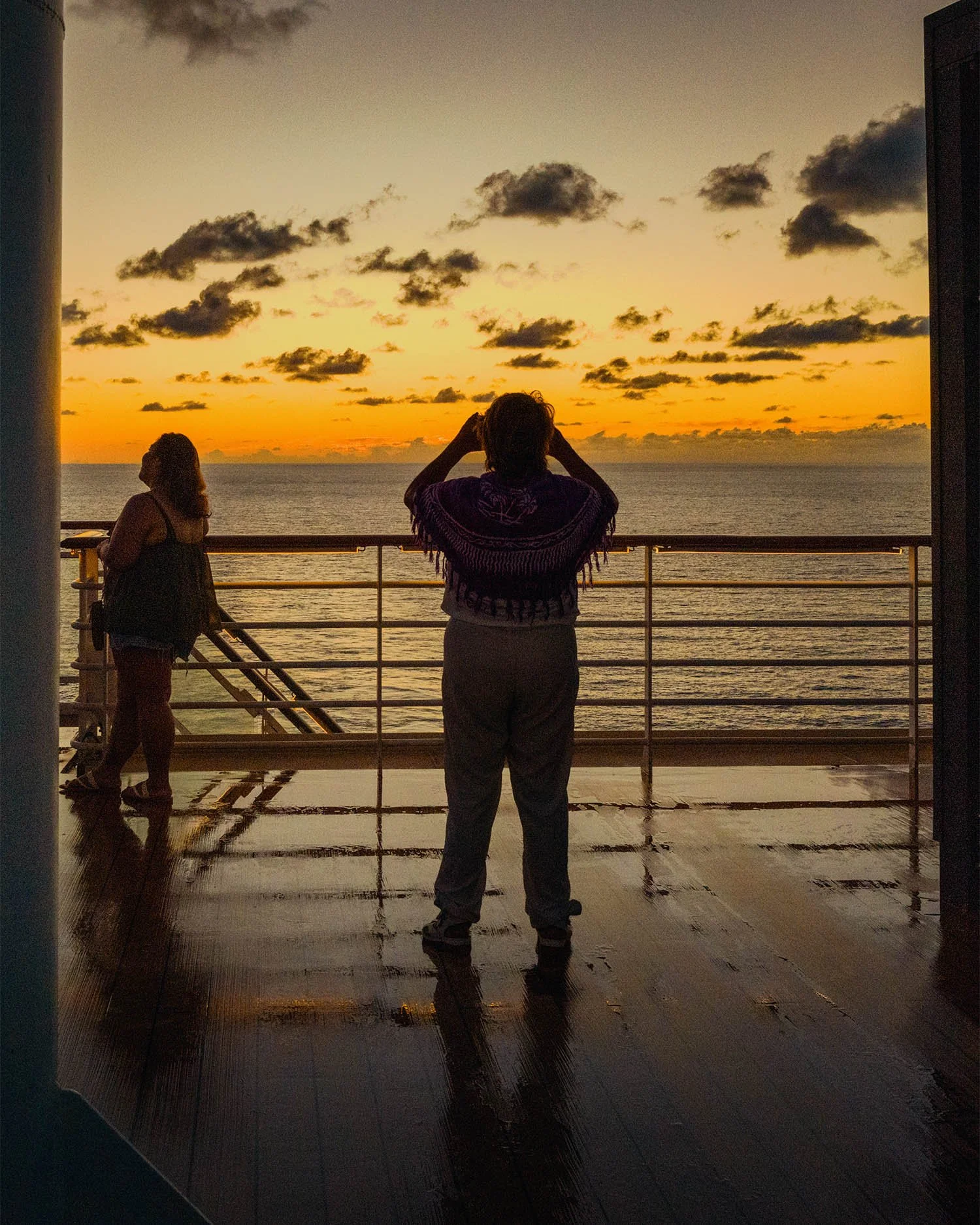 Two passengers silhouetted at ship railing during sunset, shadows and reflection on wet deck