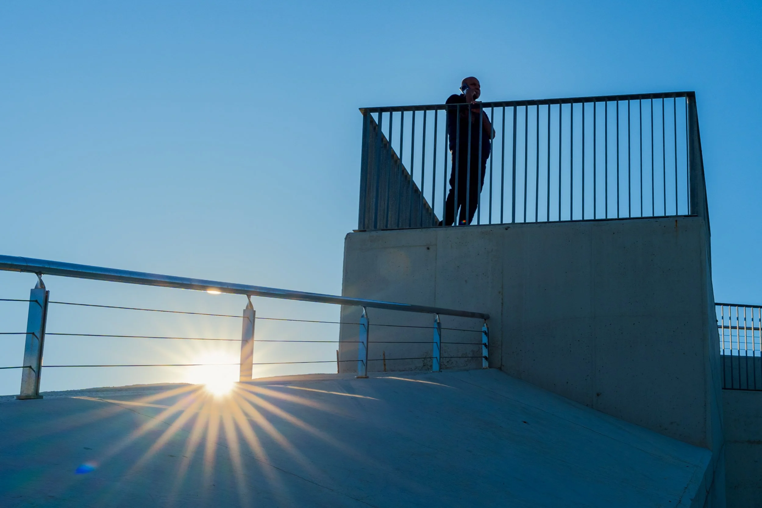 Silhouetted man on the phone leaning over a railing on a concrete platform while the sun bursts through a cable railing below