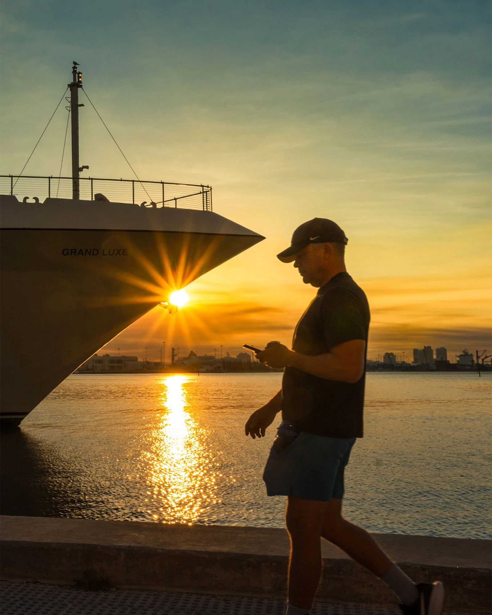 Man in a cap walking past a docked cruise ship bow with a sun flare, Bayfront Park Miami