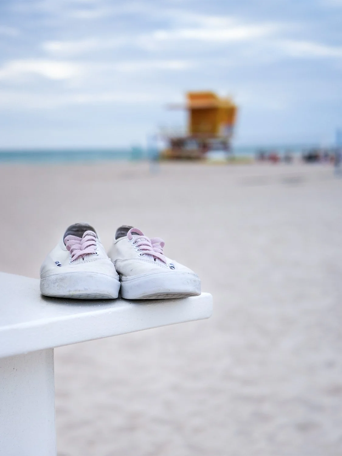 White canvas sneakers with pink laces resting on a white ledge at a sandy beach with a yellow lifeguard tower in the background