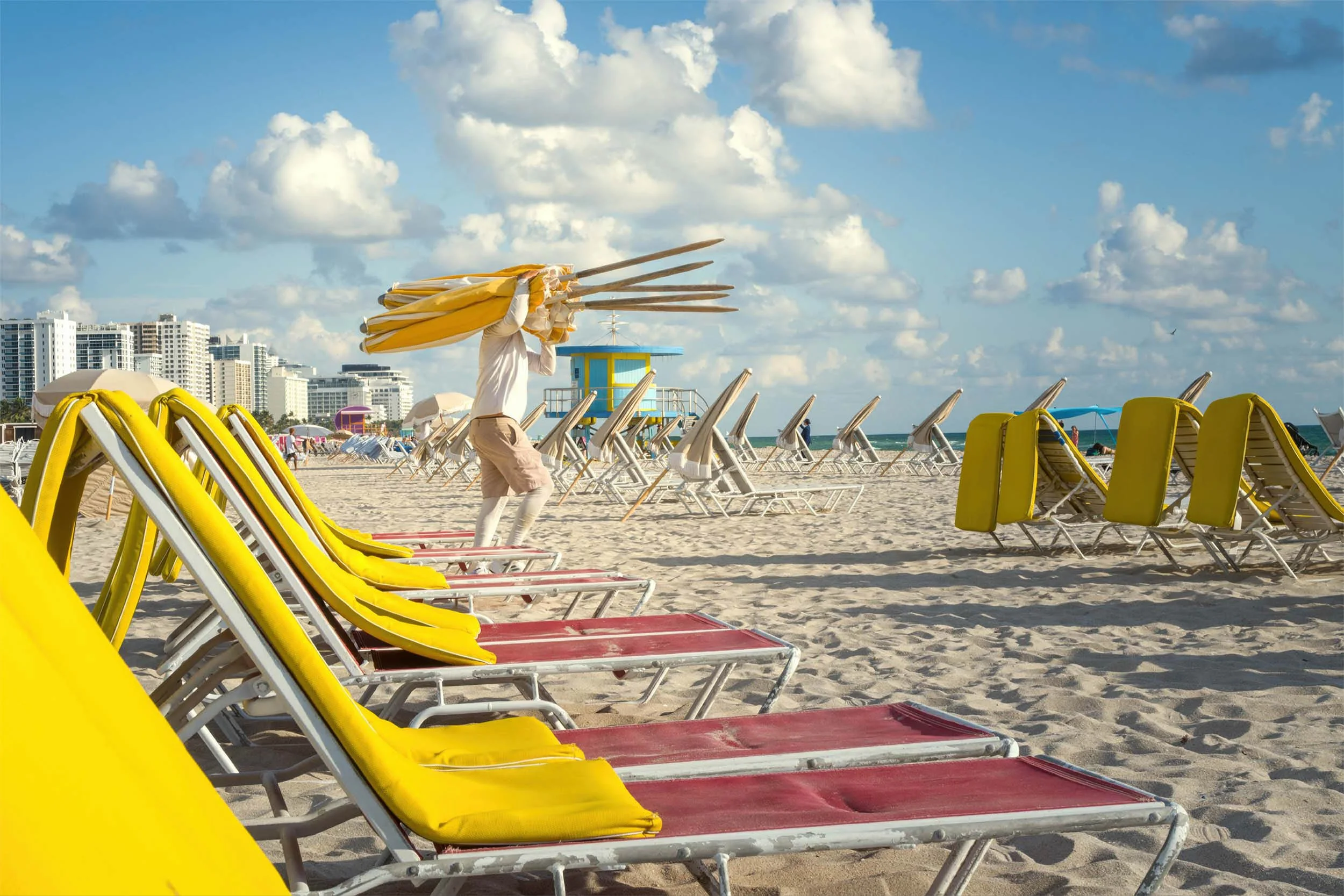 Beach worker carrying an armload of yellow umbrellas past rows of yellow lounge chairs on South Beach