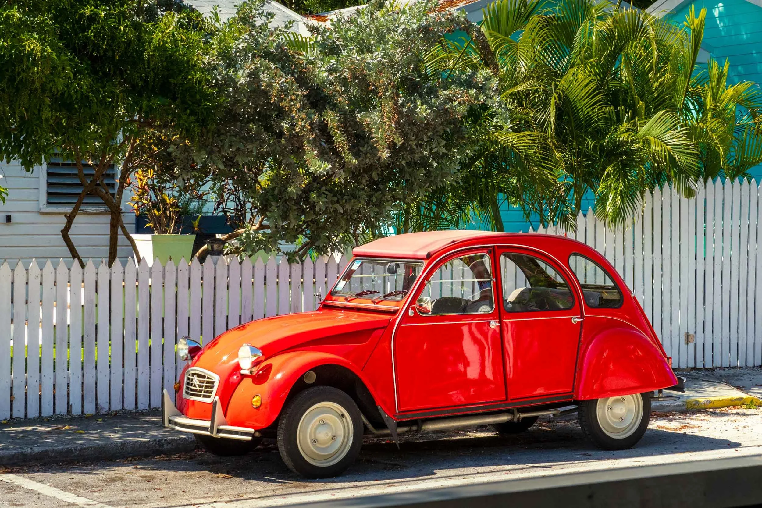 Vintage red Citroën 2CV parked on a sunny Key West street beside a white picket fence and tropical trees