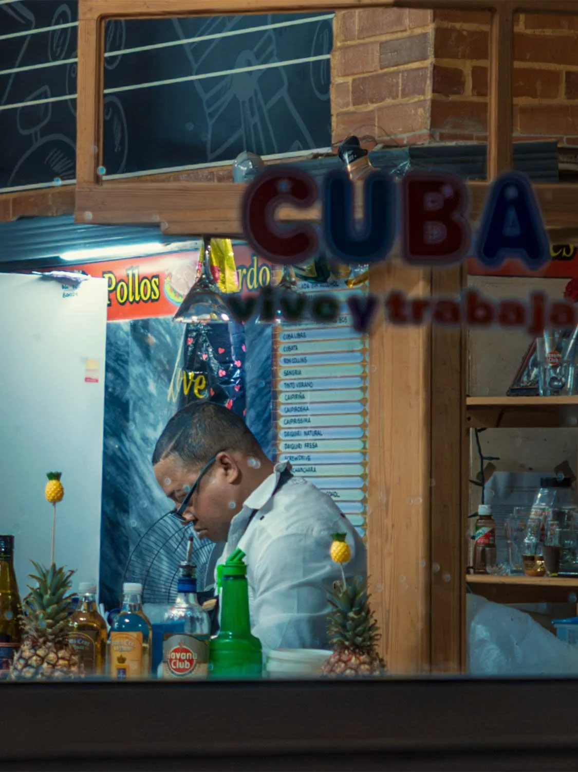 Bartender working behind a bar in Havana, seen through a window lettered "Cuba vivo y trabajo