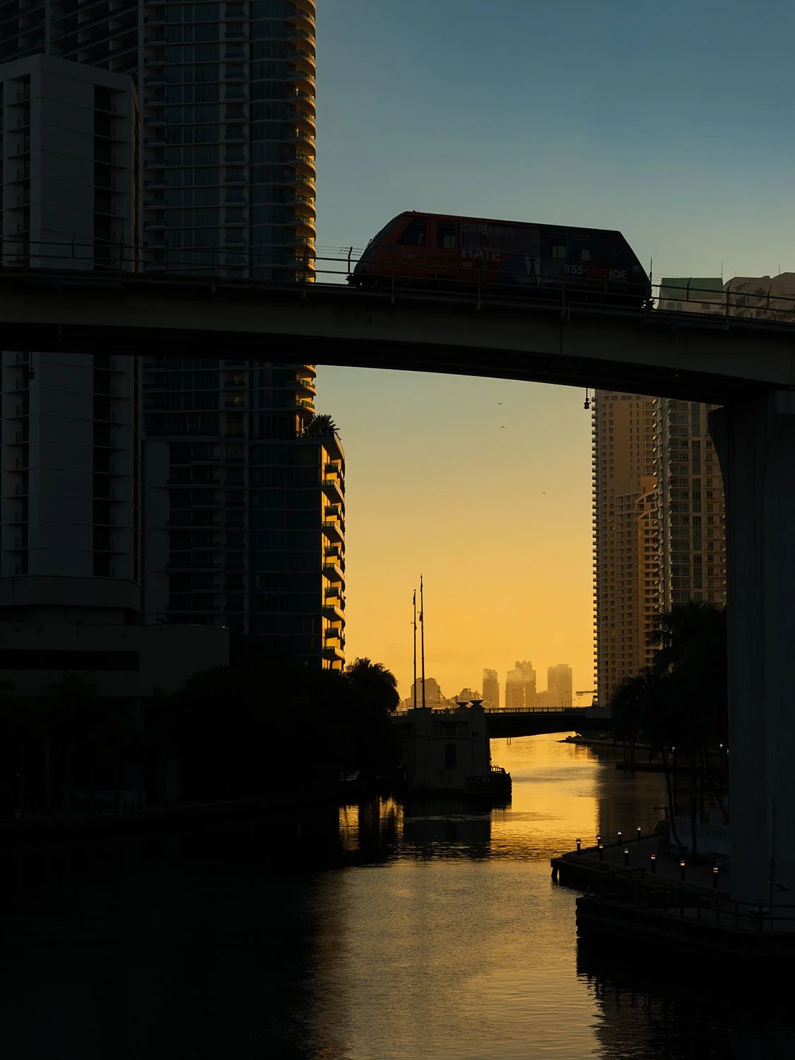 Miami River sunset with Metromover transit rail, Brickell skyscrapers, and golden sky reflection on water.