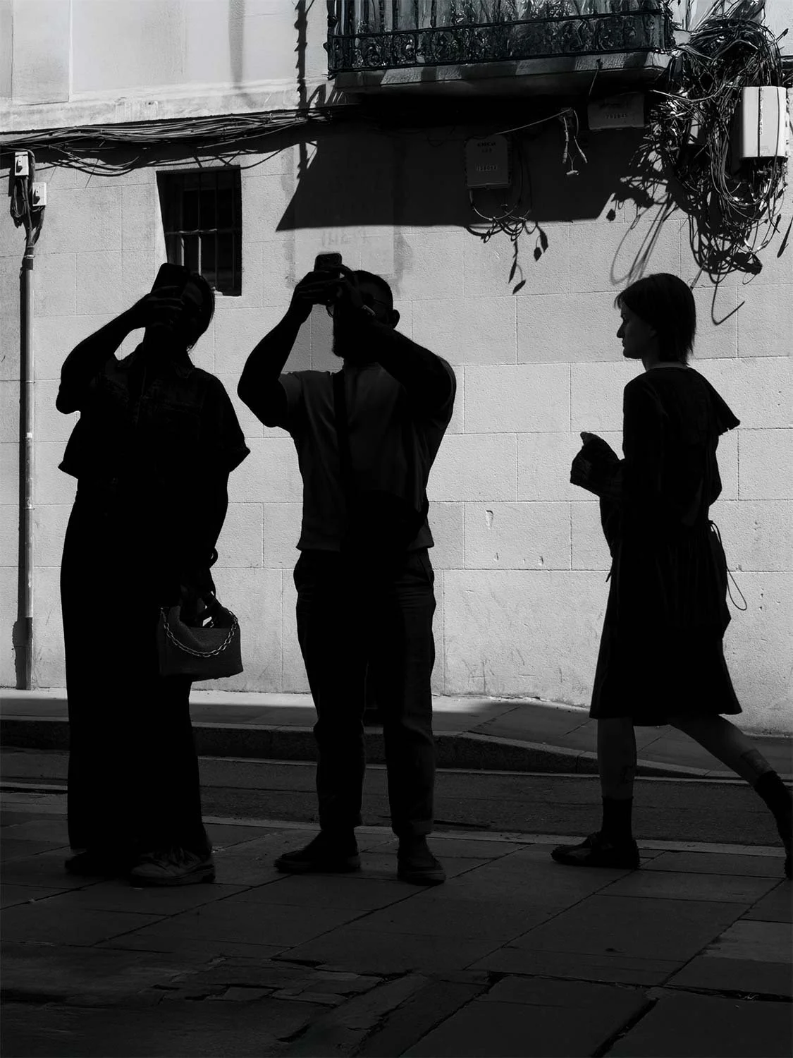 Silhouettes of two people taking photos with their phones as a woman walks past a sunlit wall in Barcelona’s Gothic Quarter