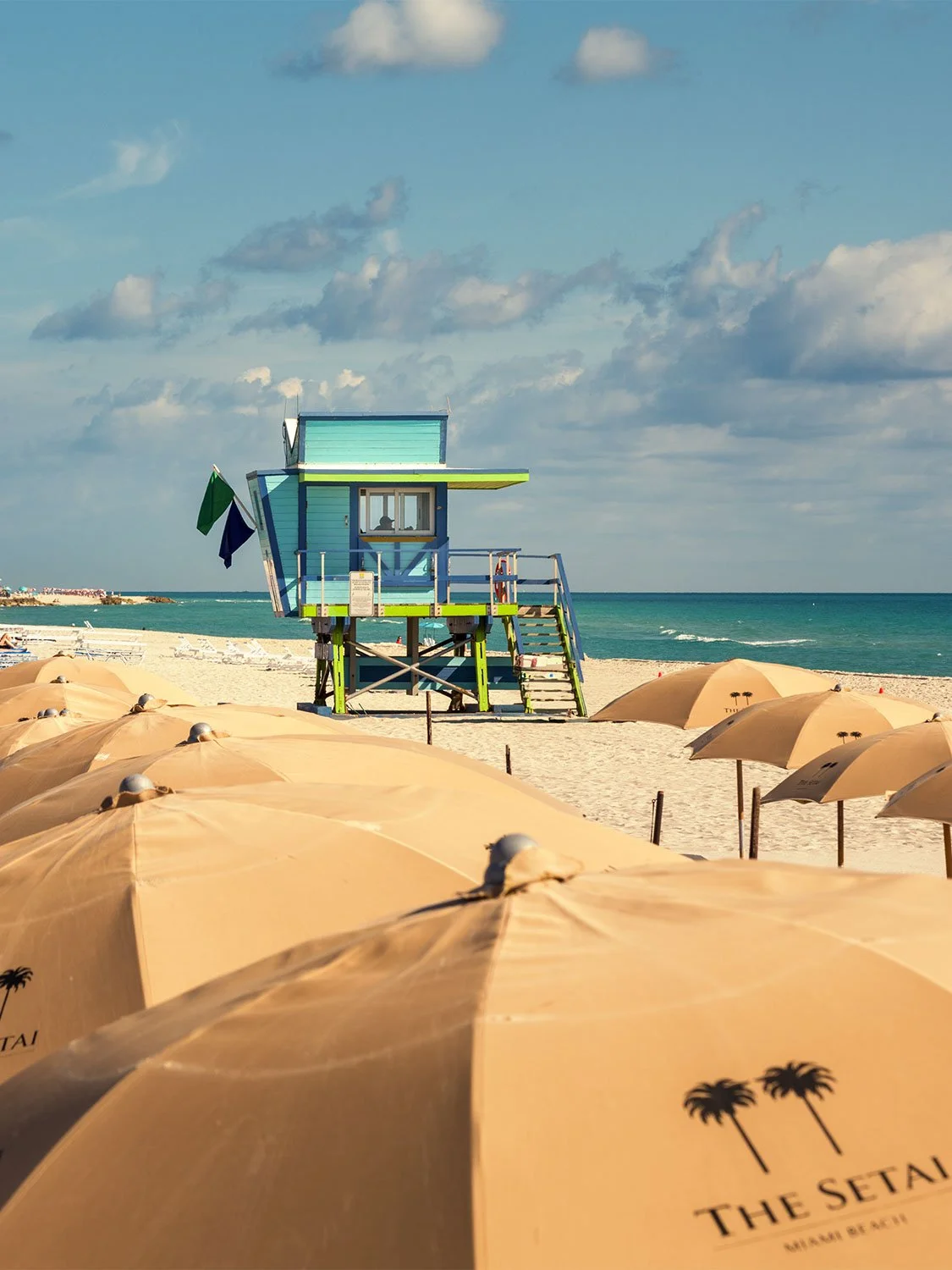 Rows of tan beach umbrellas on South Beach with a colorful lifeguard stand and turquoise ocean in the background