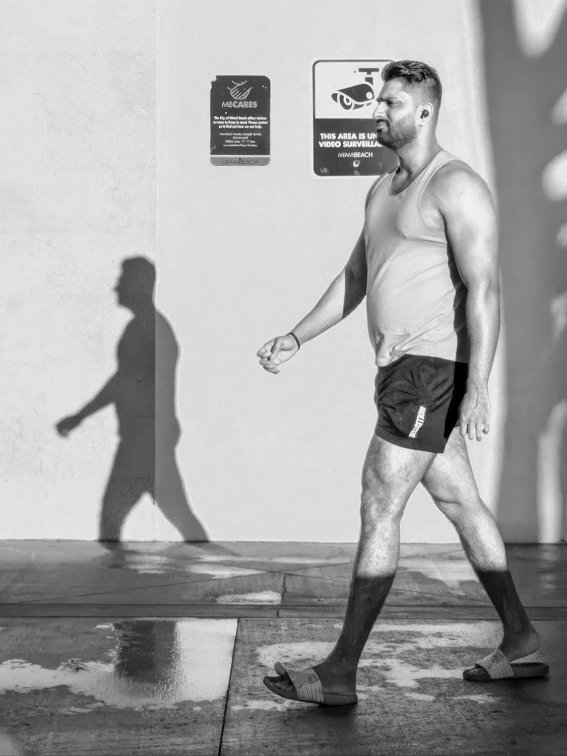 walking-shadow-bw.jpg	Man in a tank top walking beside his shadow past Miami Beach signs, in black and white