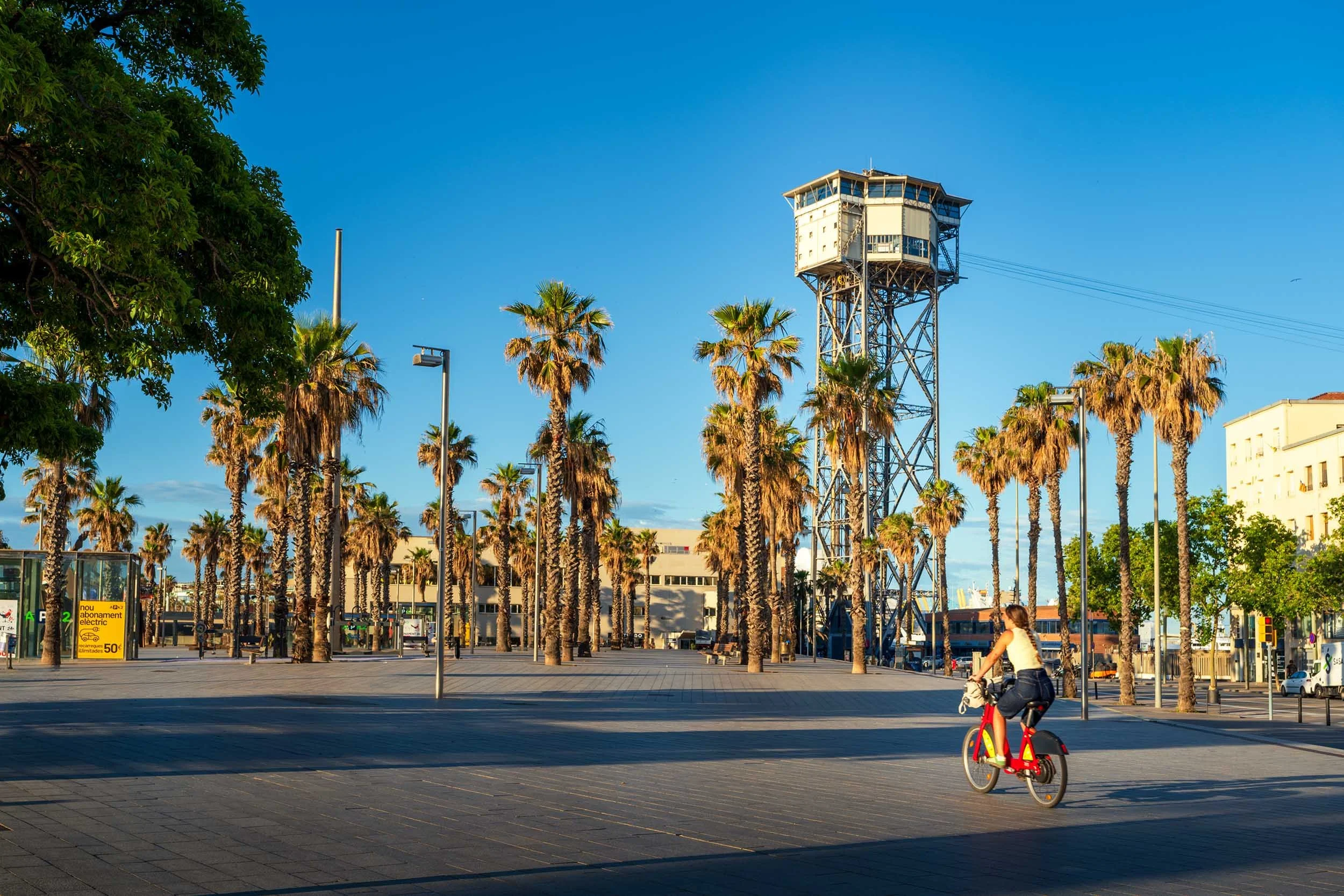Woman riding a red bike-share bicycle across a palm-lined plaza beneath the Torre de Sant Sebastià cable car tower