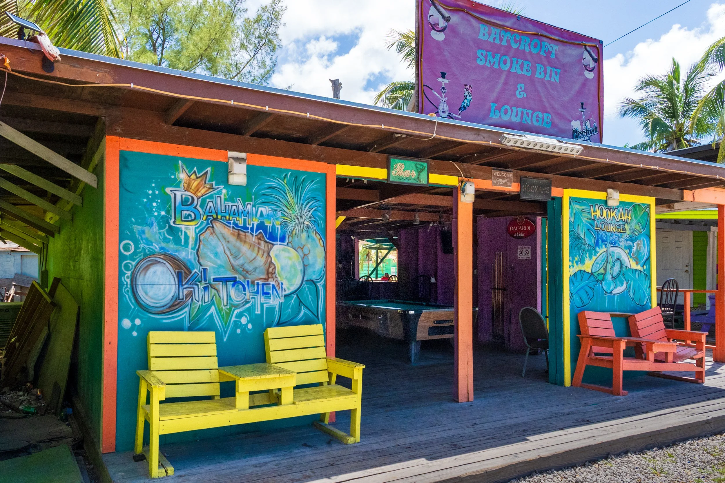 Colorful open-air bar with Bahamian Kitchen and Hookah Lounge murals, yellow bench, pool table, at Arawak Cay Nassau.