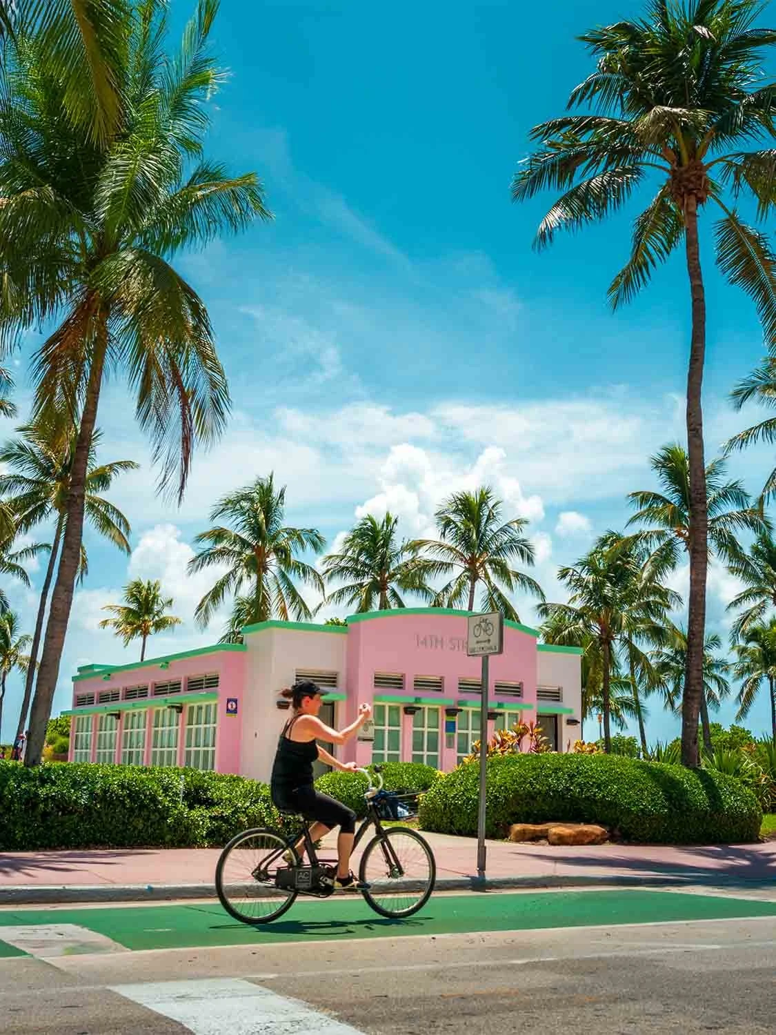 Woman cycling past a pink Art Deco building on a green bike lane in Miami Beach, framed by tall palm trees