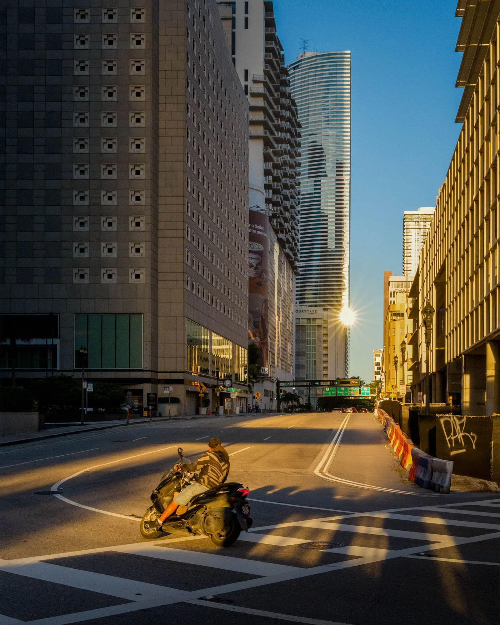 Scooter rider crossing an intersection with a sun flare at the building edge, Biscayne Blvd and SE 3rd St, downtown Miami