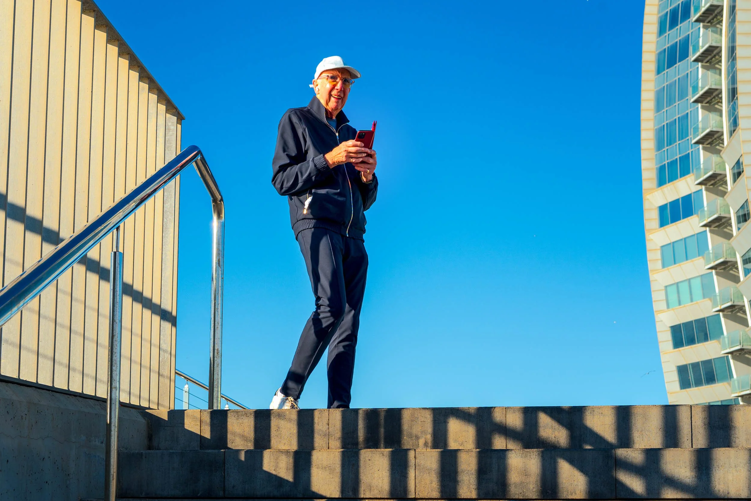 Smiling older man in a white cap and navy tracksuit holding a red phone at the top of concrete steps in Barceloneta, a modern tower against the blue sky
