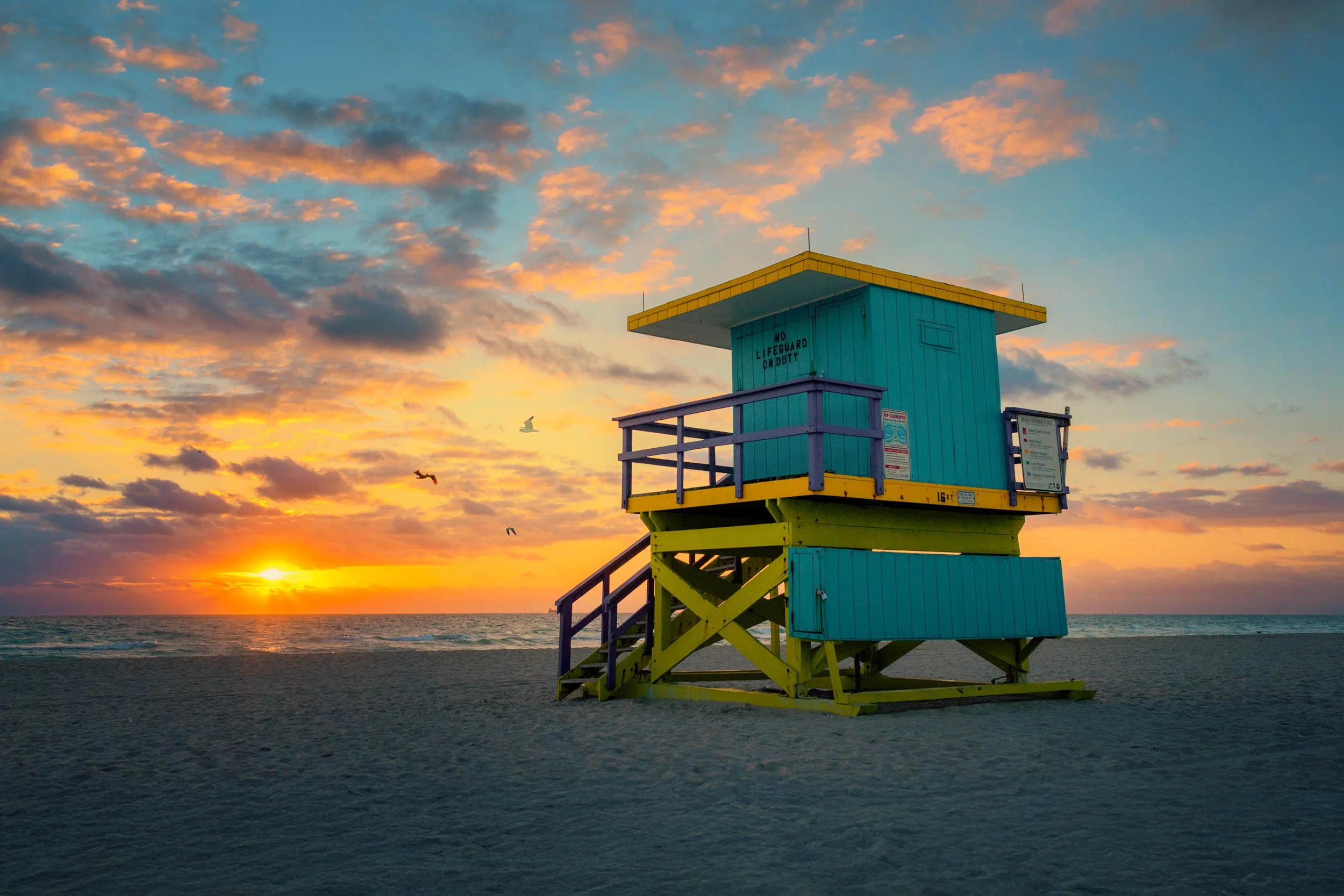 A teal and yellow lifeguard tower at 16th Street against a vivid sunrise sky with orange and purple clouds, as birds fly over the ocean on South Beach.