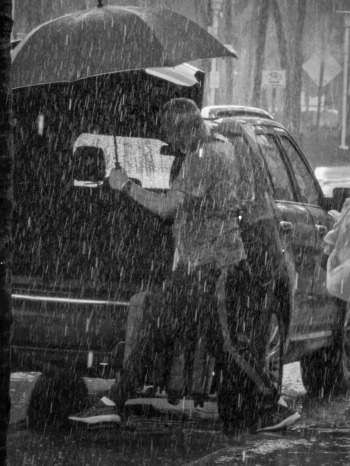 Person holding umbrella in heavy rain beside a parked SUV on a wet city street, black and white photo