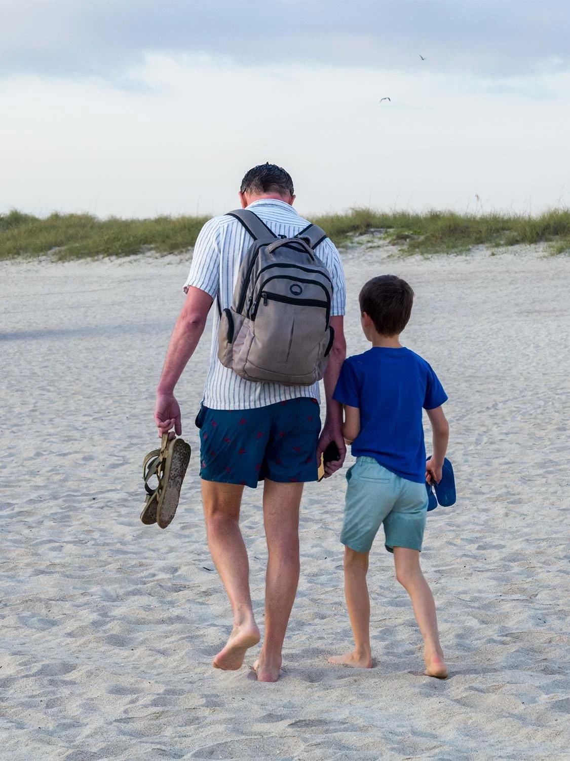 Adult and child walking barefoot on a sandy beach toward dunes, both carrying sandals, viewed from behind