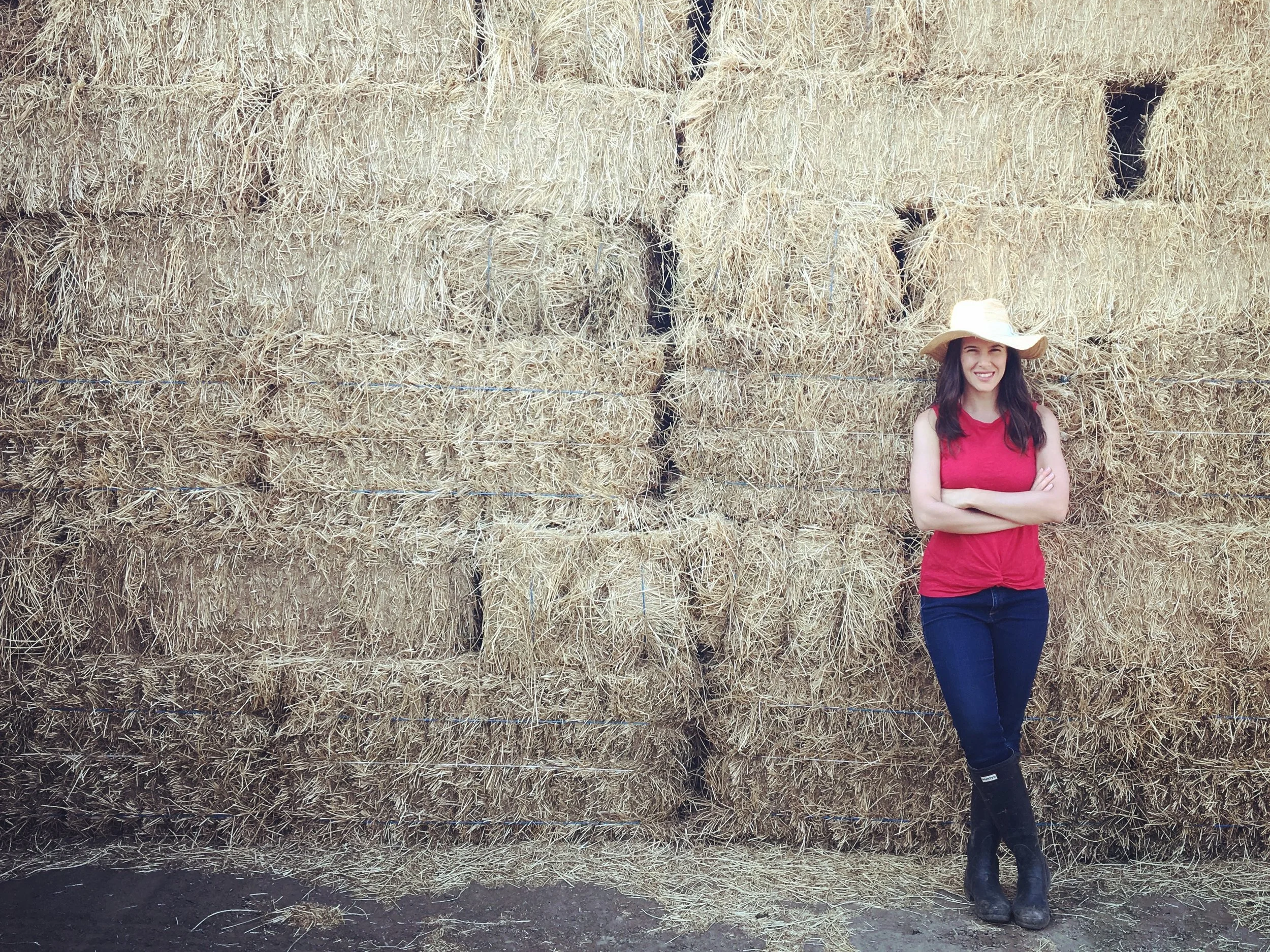 A woman, Nicole Jolly, in a red sleeveless top, jeans, and black boots standing with arms crossed in front of a large wall of stacked hay bales, wearing a wide-brimmed straw hat. During a film shoot in California for How Does it Grow?