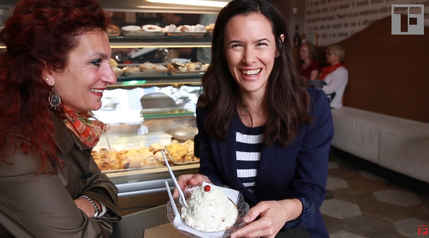 A woman, Nicole Jolly, smiling, seated with another woman in a cafe in Naples, Italy, holding a plate with a lemon dessert topped with whipped cream and a cherry.