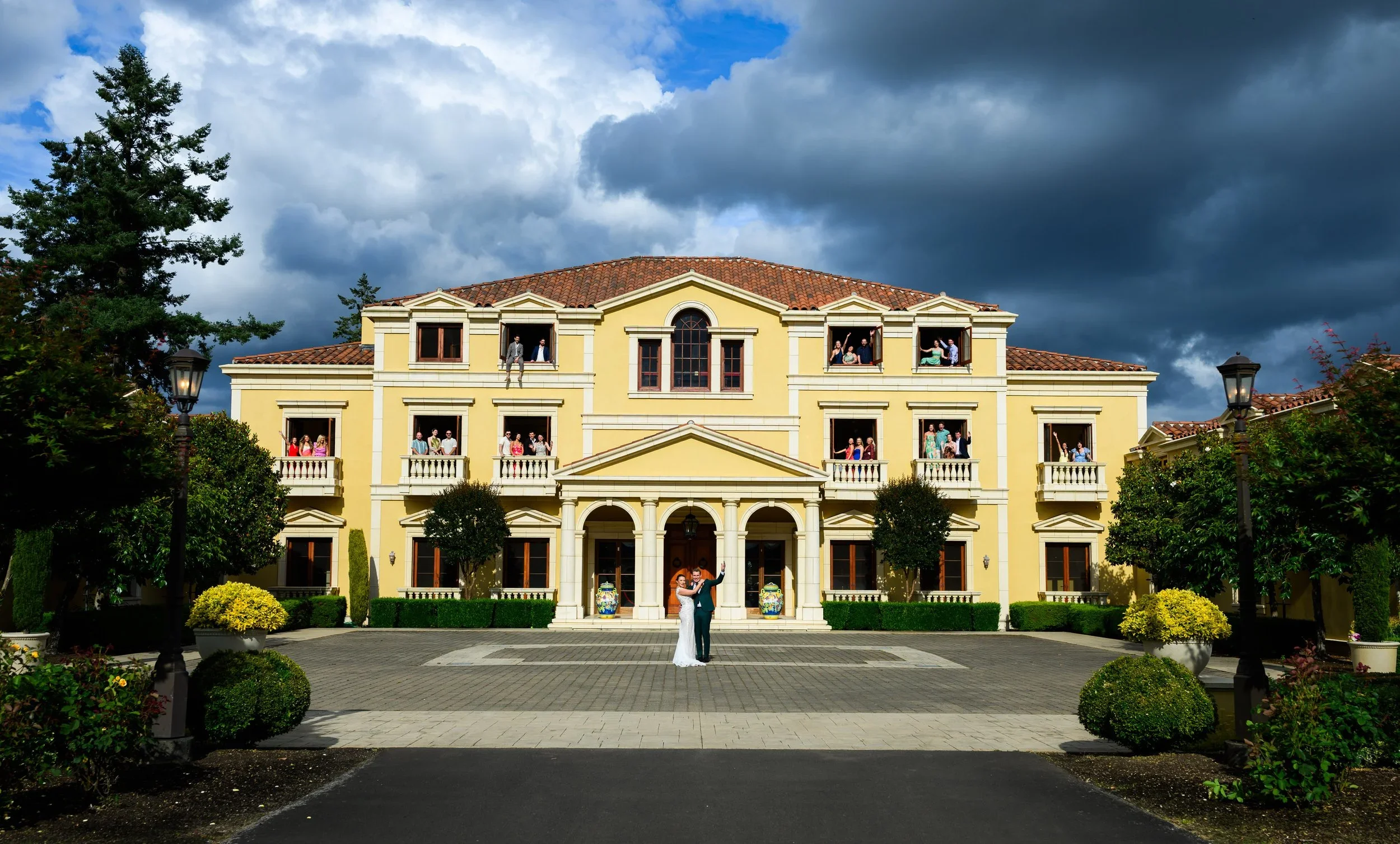 A wedding couple standing in front of a large yellow mansion, with guests seen in the windows above, and dark storm clouds in the sky.