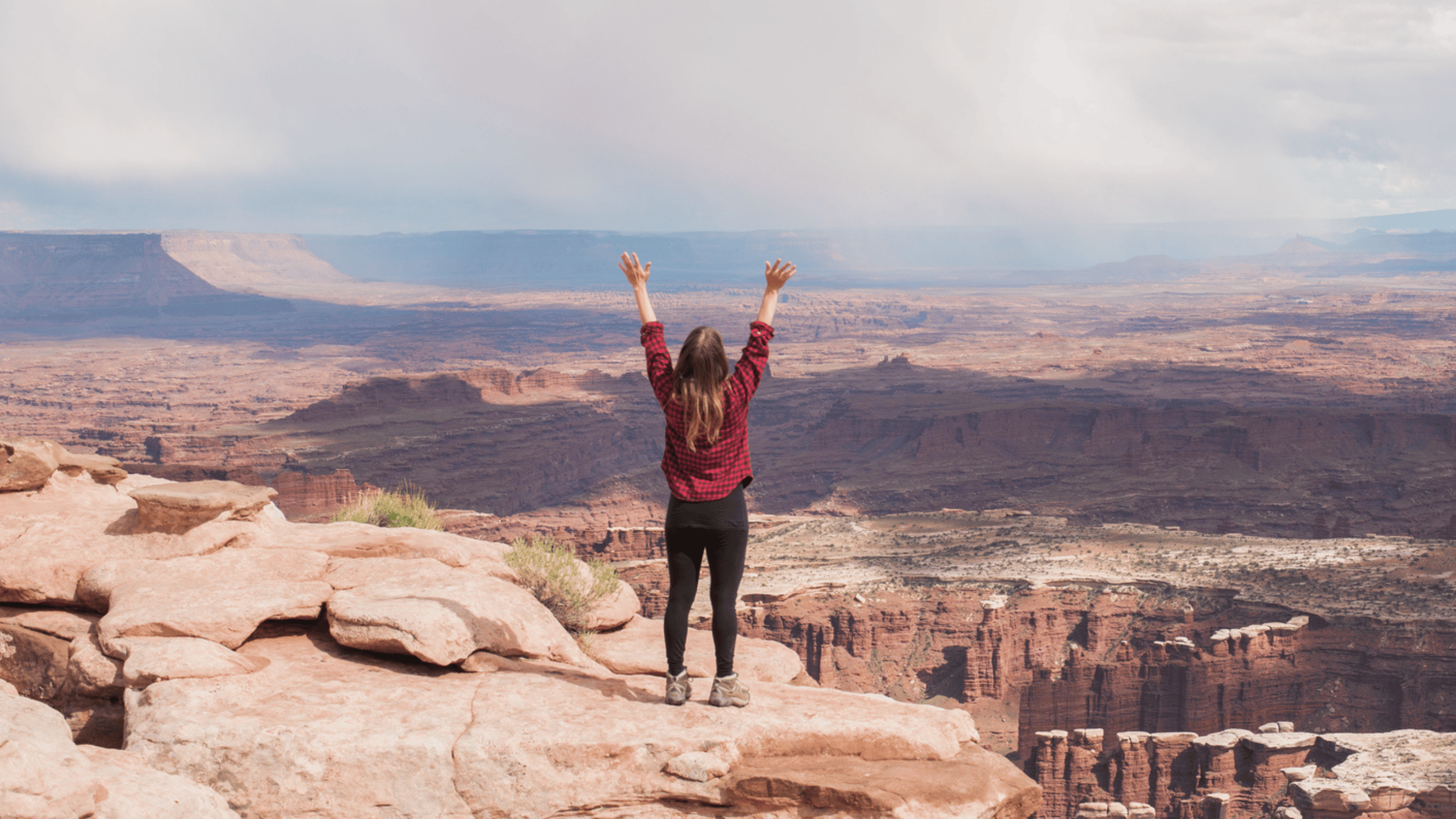 A women reaches her hands up to the sky from the top of a cliff overlooking a beautiful desert landscape