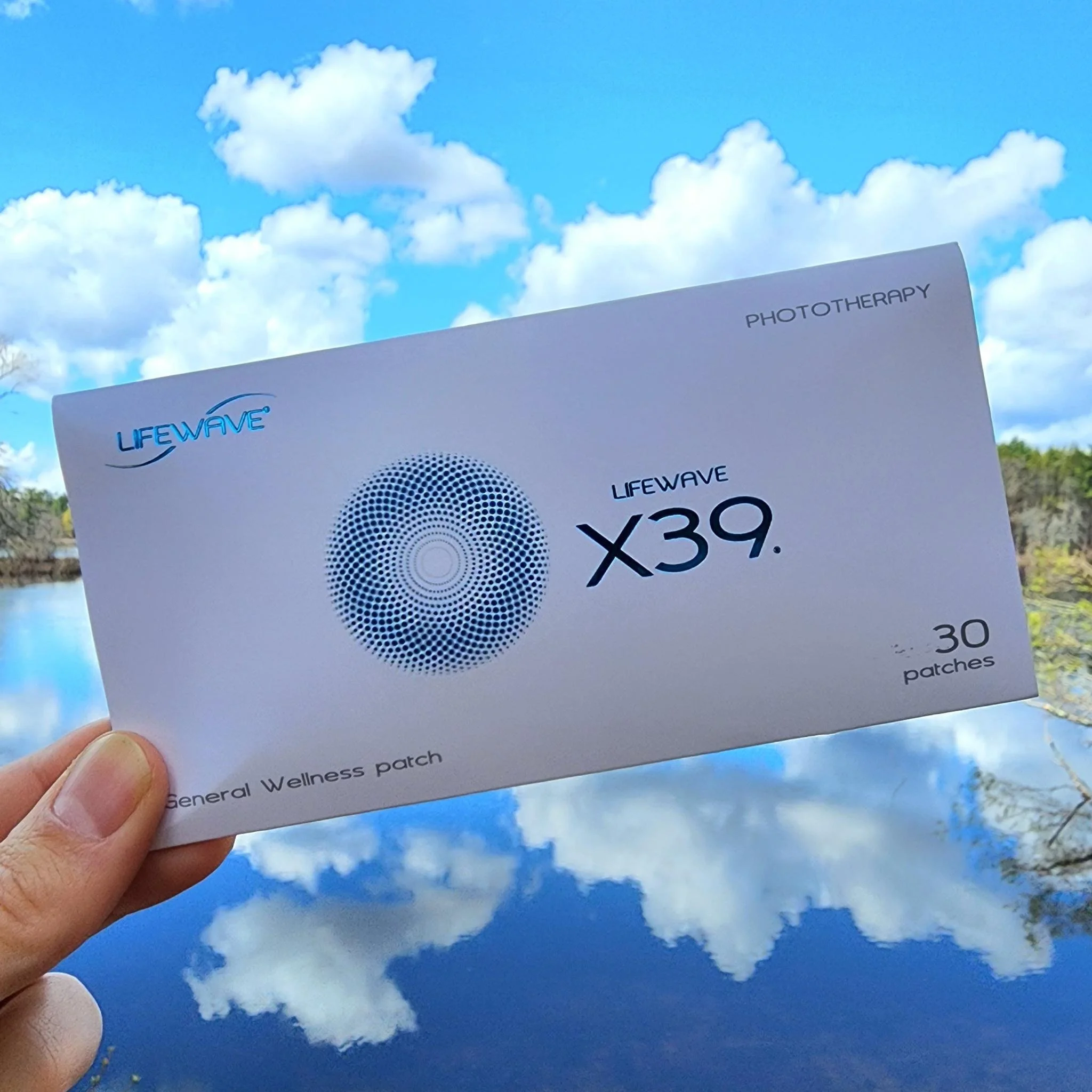 A hand holding a pack of LifeWave X39 phototherapy patches in front of a nature background with clouds, sky, and water reflections