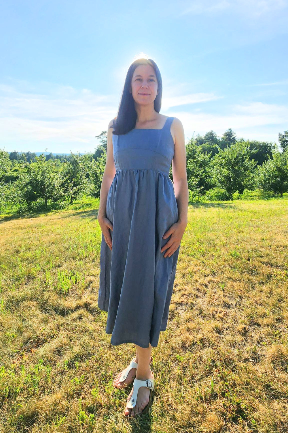 Kara is wearing the light blue 100% European Linen Fit & Flare Midi Dress by Quince standing on a green hill in front of an orchard