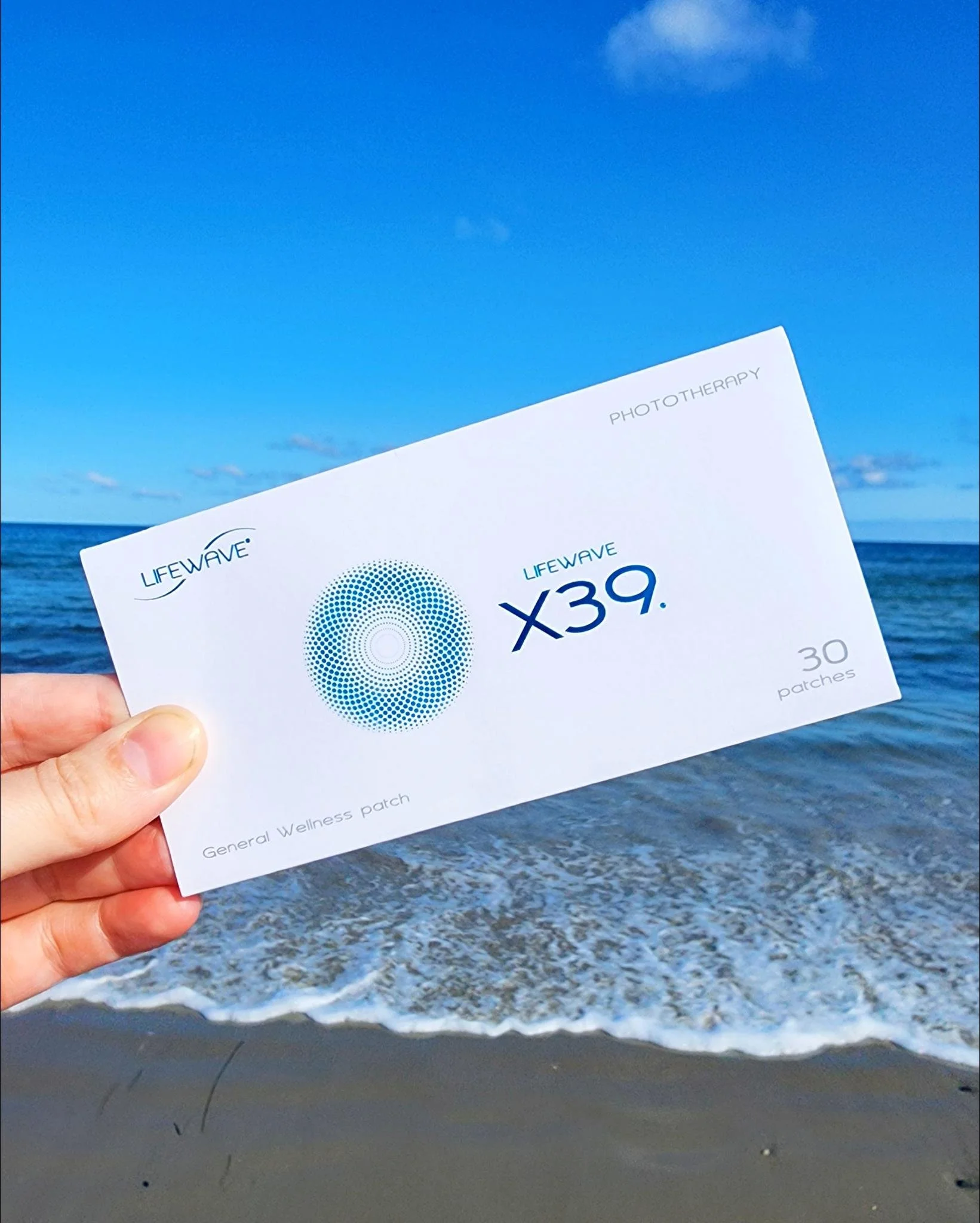 Hand holding a LifeWave X39 Patch Pack in front of ocean waves and a blue sky at the beach