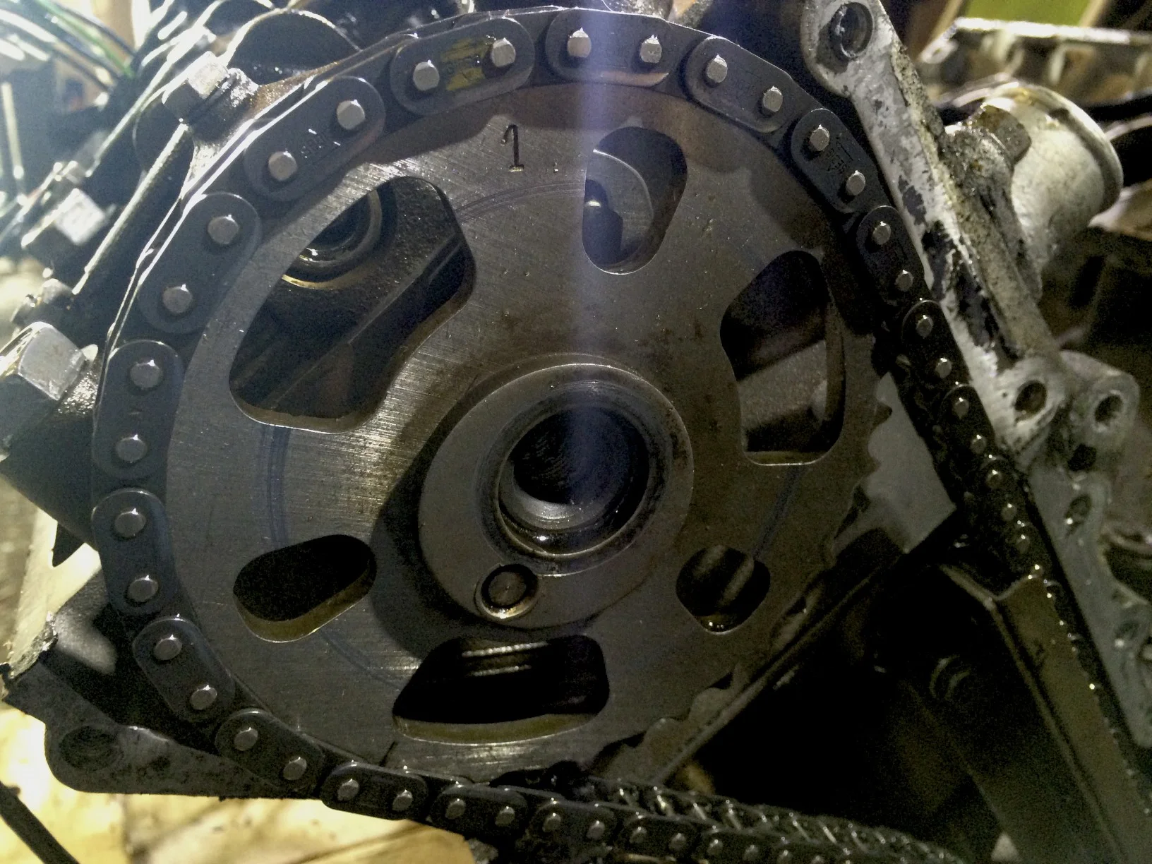 The timing chain on the sprocket, but with the hex screw removed. The '1' at the top of the sprocket lines up with the yellow mark on the timing chain when the engine is properly timed, The pin below the center hole lines up with a recess in the cams and keeps the sprocket locked with the cams' rotation.
