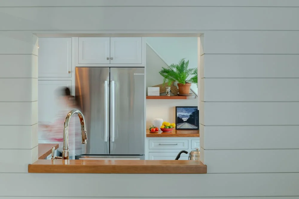 View of a modern kitchen through a rectangular opening in a white shiplap wall, featuring a stainless steel refrigerator, a wooden countertop with a sink, a potted plant, and a small monitor.
