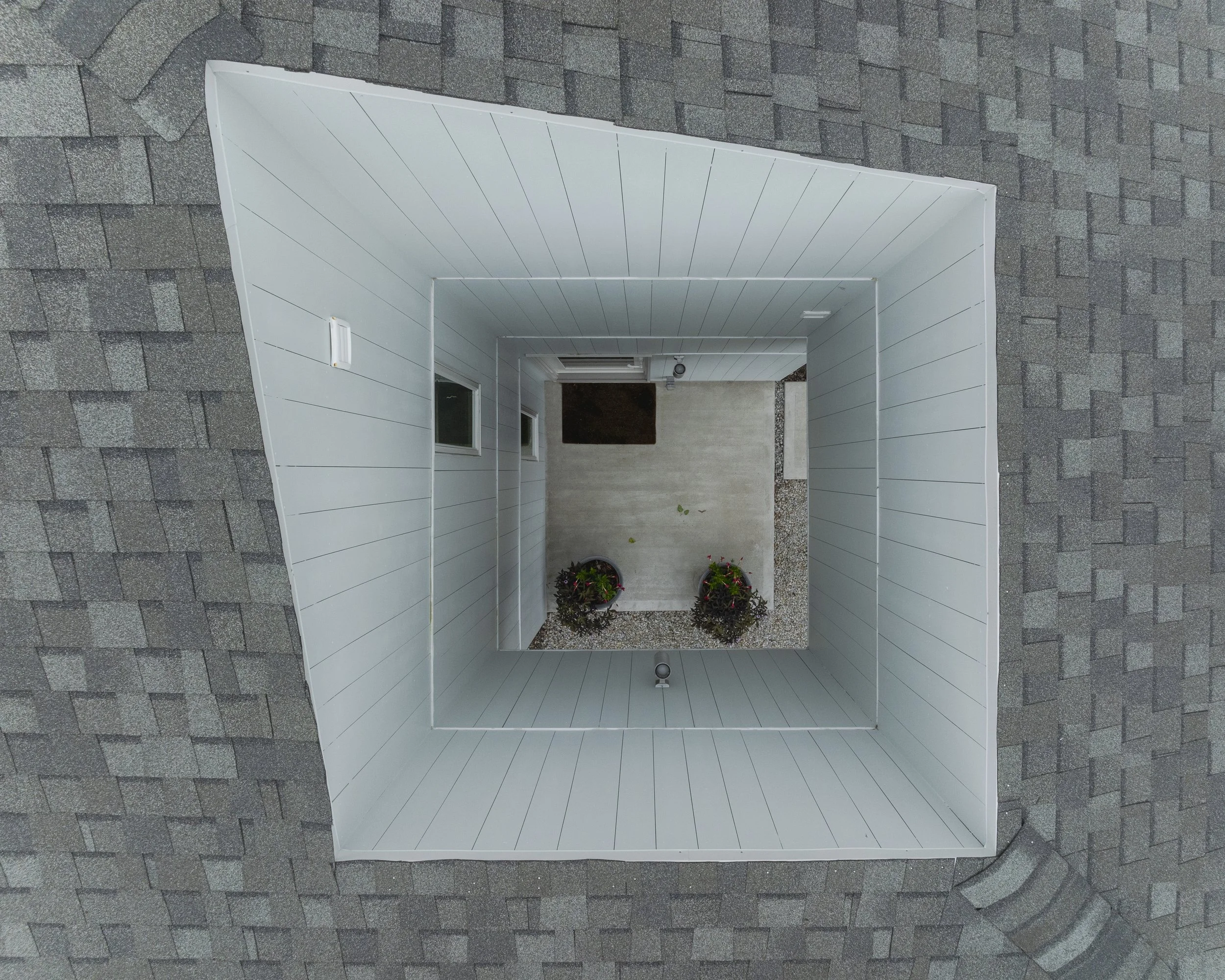 A view from above of a small outdoor area with white fencing, potted plants, a doormat, a door, and windows, surrounded by gray roofing shingles.