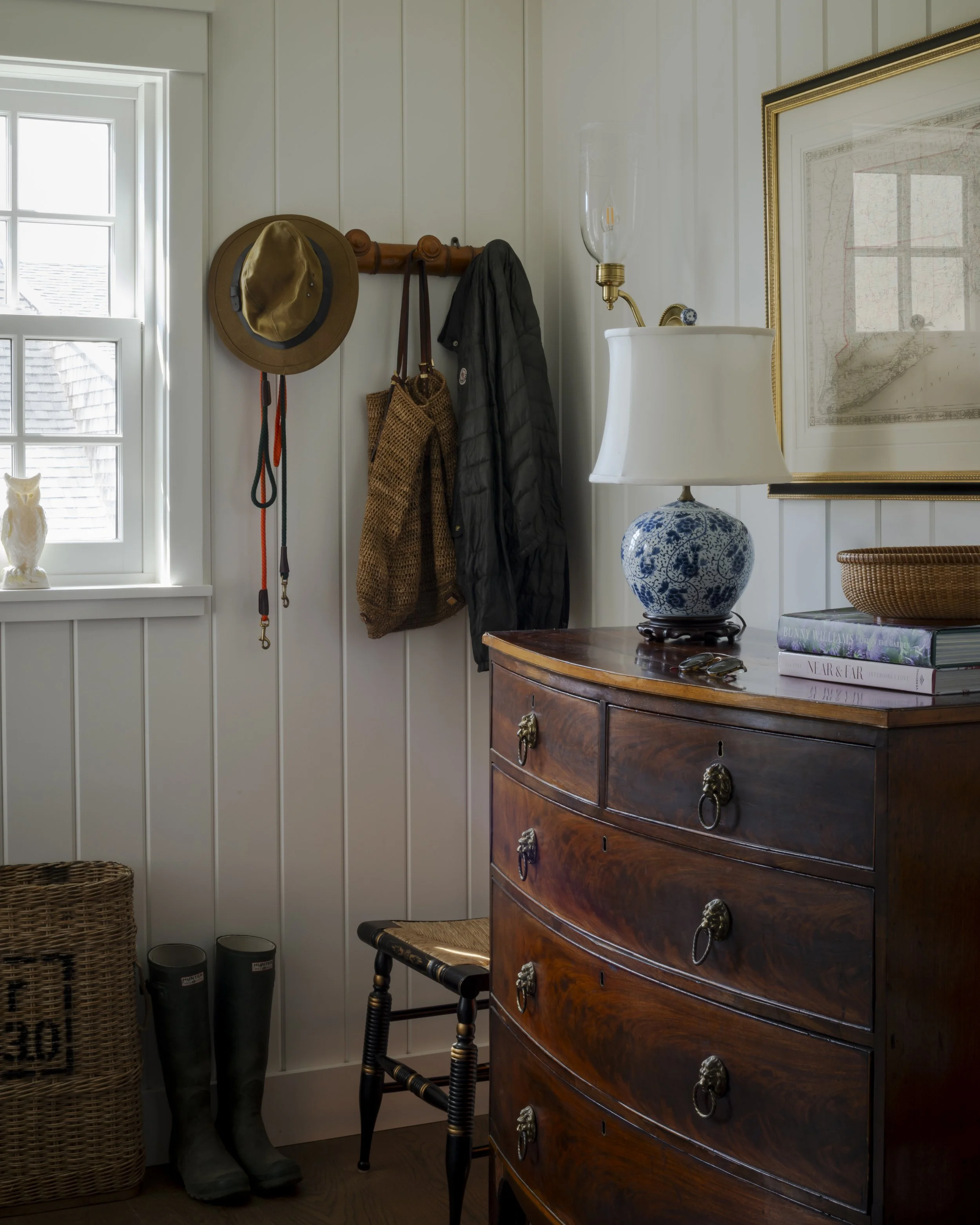 An entryway area with a window, a coat rack with a hat and coats, a wicker basket, rain boots, a wooden dresser with a lamp, books, and a framed map on the wall.