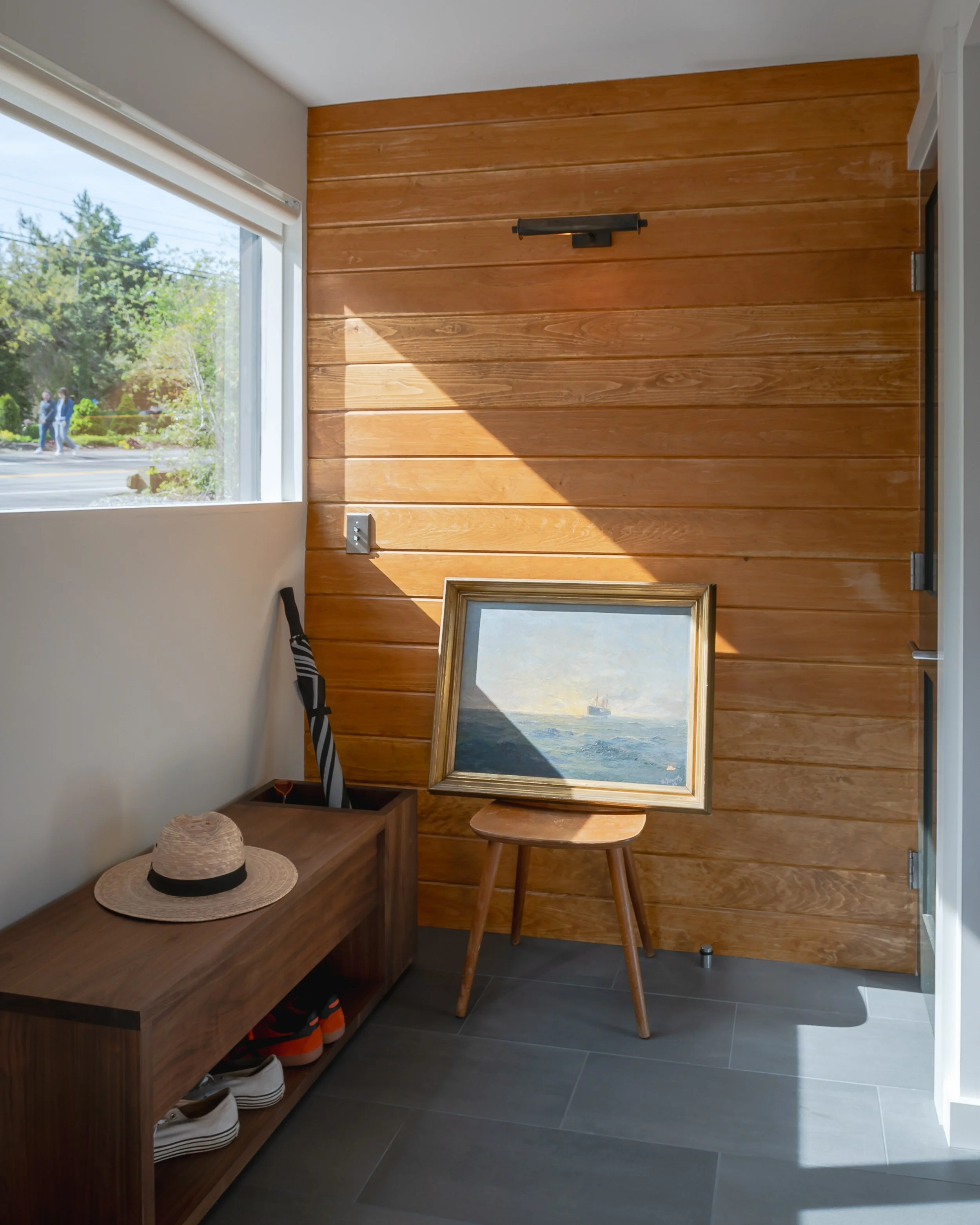 Interior of a room with a wooden wall, a window, a framed painting of a seascape on a stool, a wooden shoe rack with shoes, a straw hat, and an umbrella, with sunlight casting shadows.