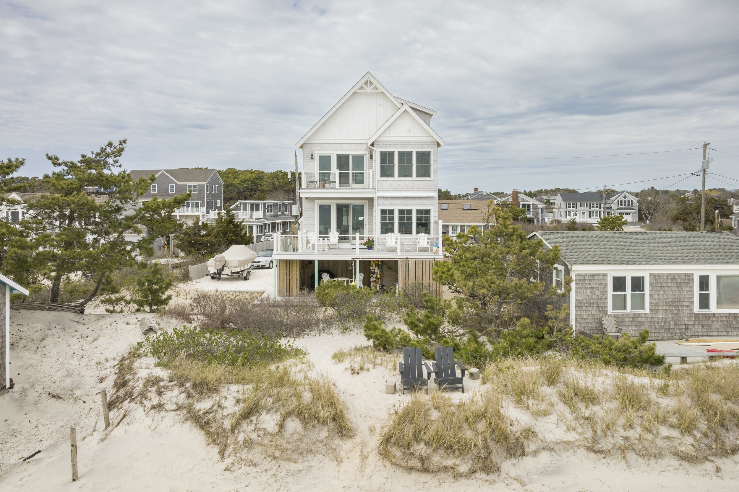 Beach house with multiple decks, surrounded by sand dunes and beach grass, and neighboring houses in the background under cloudy sky.