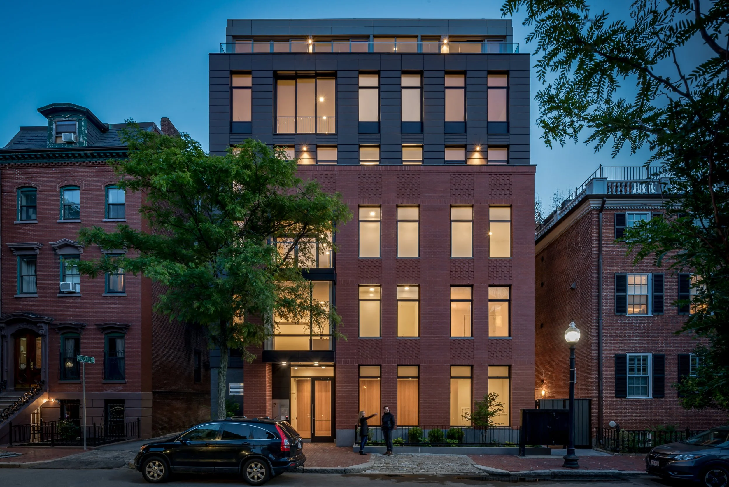 A modern multi-story building with a brick facade on a city street at dusk, with trees, parked cars, and two people talking outside.