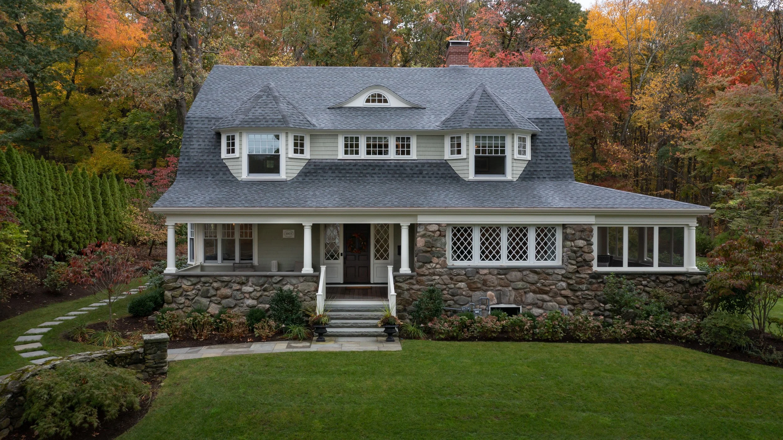 A large, two-story house with a stone foundation, white siding, and a dark gray shingled roof surrounded by trees with fall foliage.