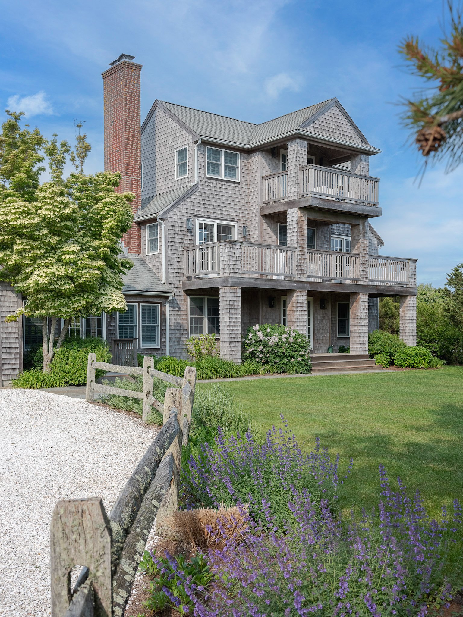 A multi-story house with gray shingle siding, multiple balconies, and a red brick chimney, surrounded by green trees and bushes, with a gravel path and purple flowers in the foreground.