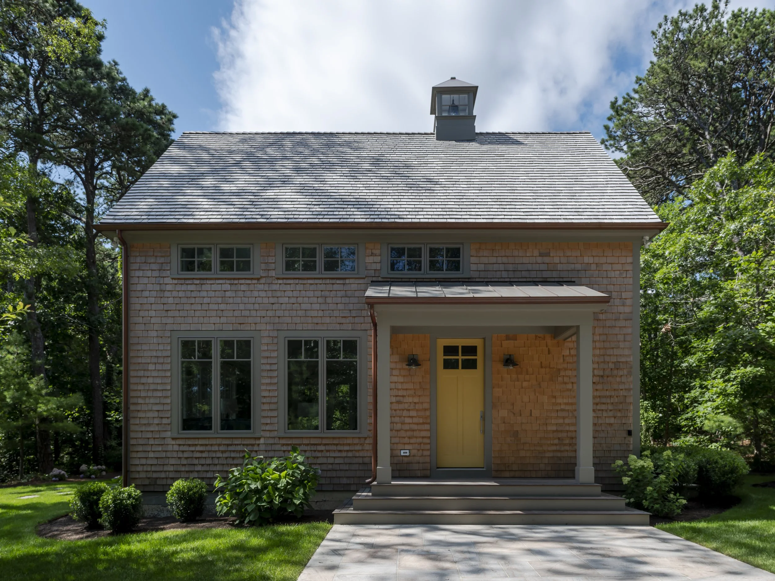 Front view of a two-story house with wooden shingle siding, a yellow front door, gray framing, and a gabled roof, surrounded by green trees and a manicured lawn.