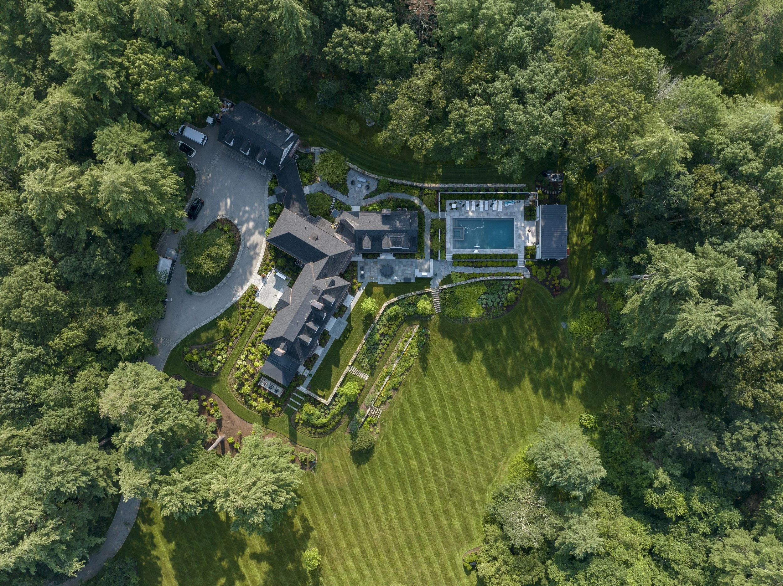 An aerial view of a large house with a driveway, surrounded by lush green trees, a well-maintained lawn, a swimming pool, and landscaped gardens.