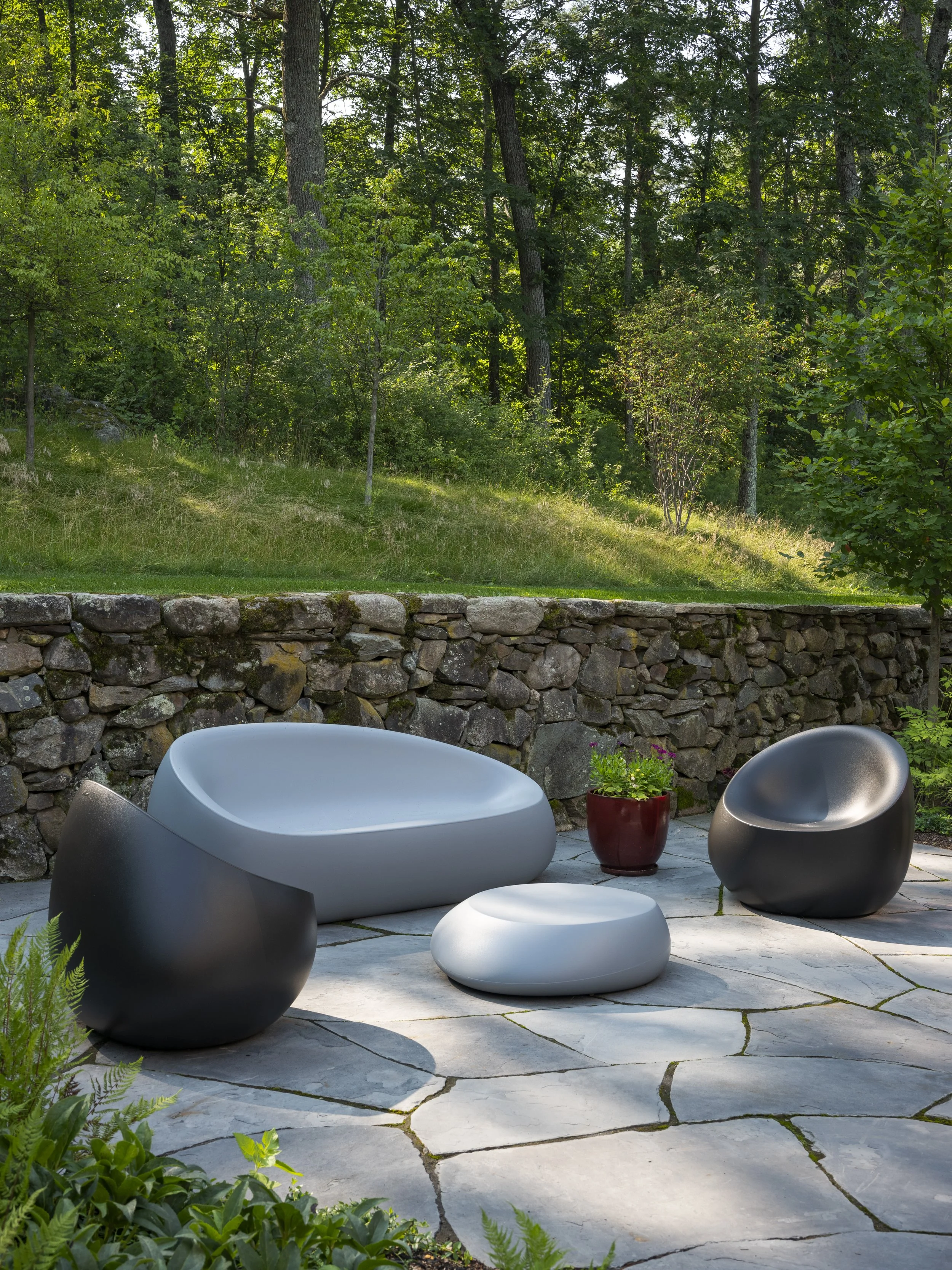 Outdoor patio with modern gray and black lounge chairs, a small round gray table, a red flower pot, and a stone wall, set against a lush green forested landscape.