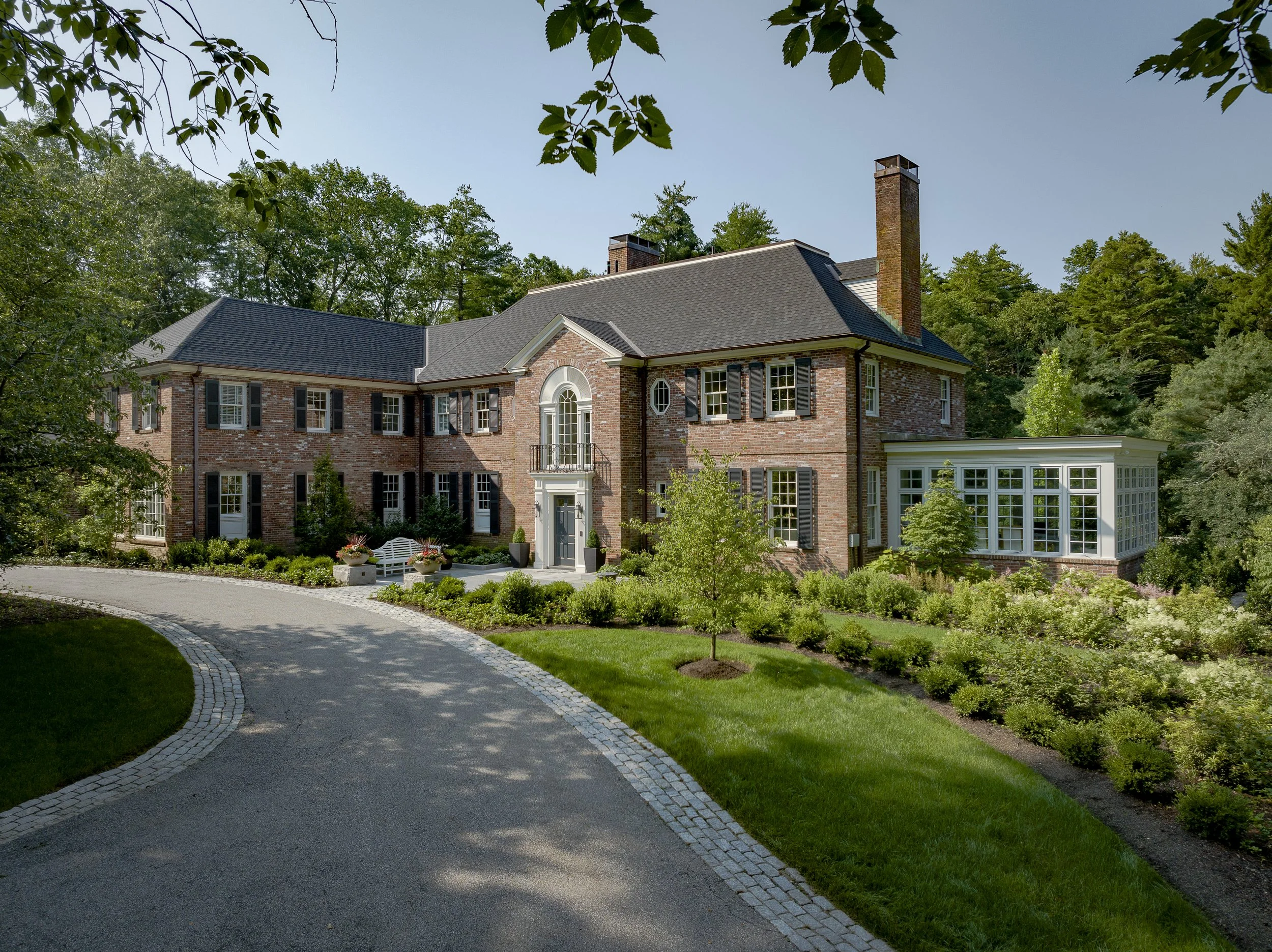 Large brick house with multiple chimneys, black shutters, and a sunroom with lots of windows, surrounded by lush green gardens and a curved driveway.
