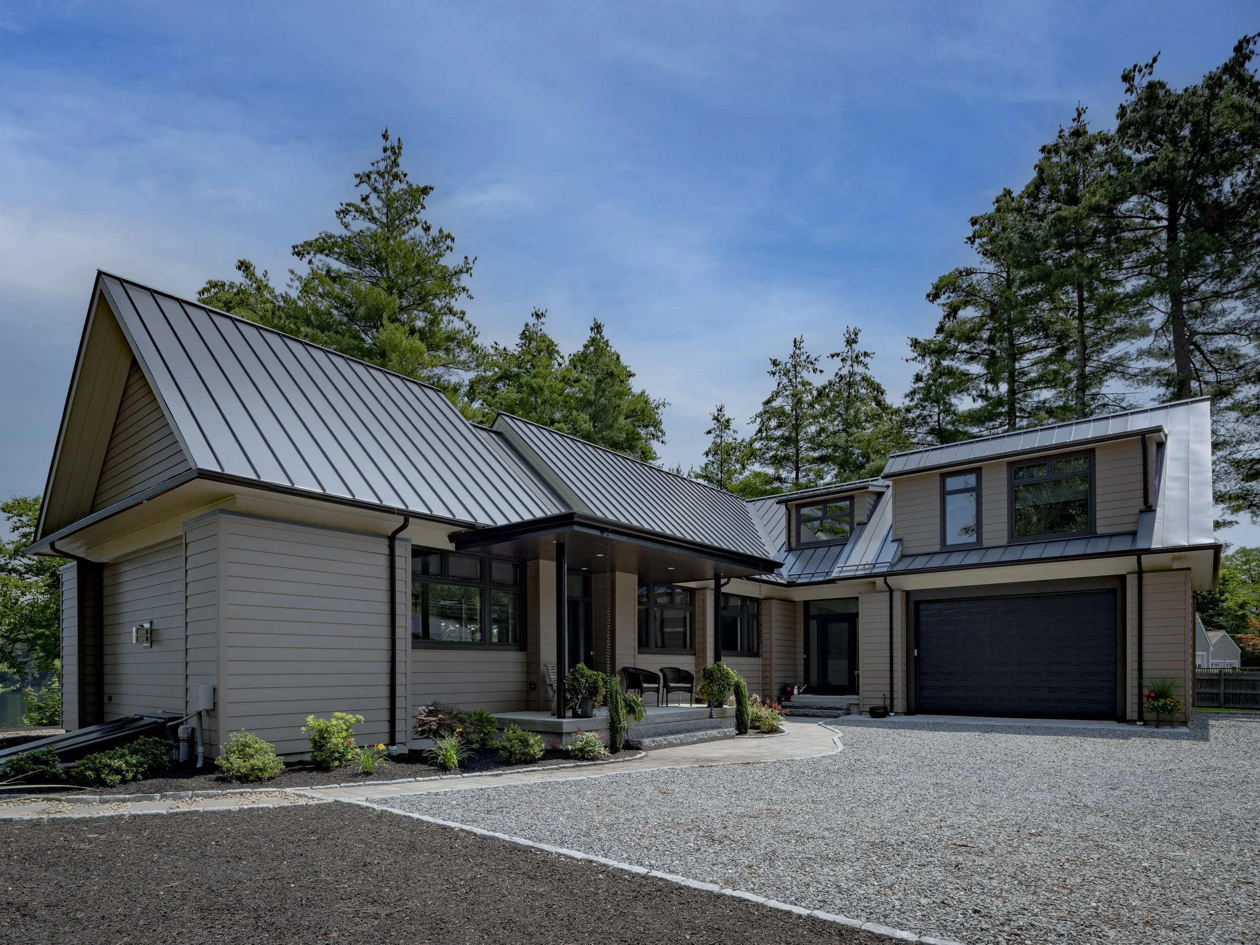 Modern house with a metal roof and black garage door, surrounded by trees and greenery, on a gravel driveway.