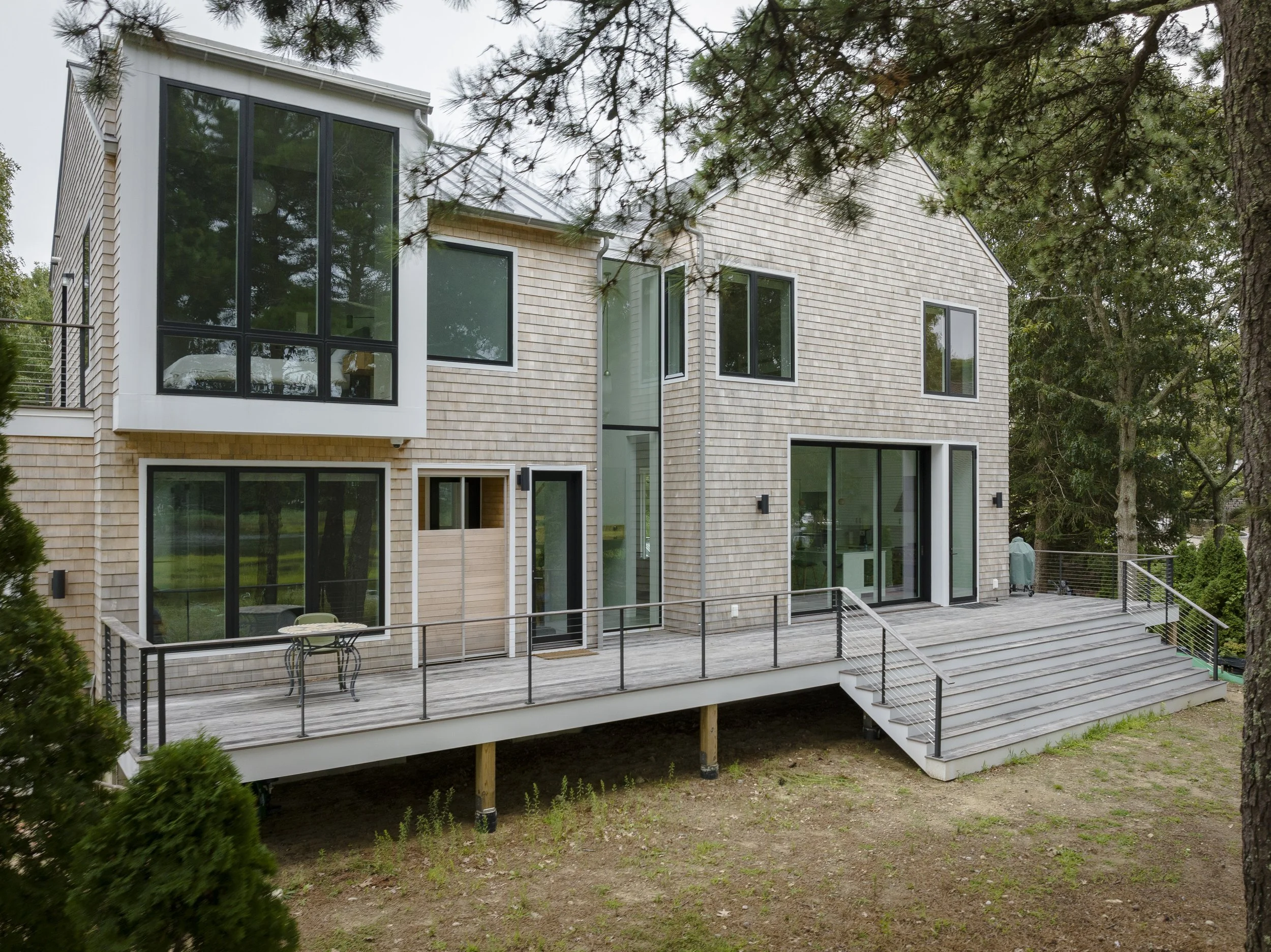 Modern two-story house with a large wooden deck, glass double doors, and multiple windows, surrounded by trees and greenery.