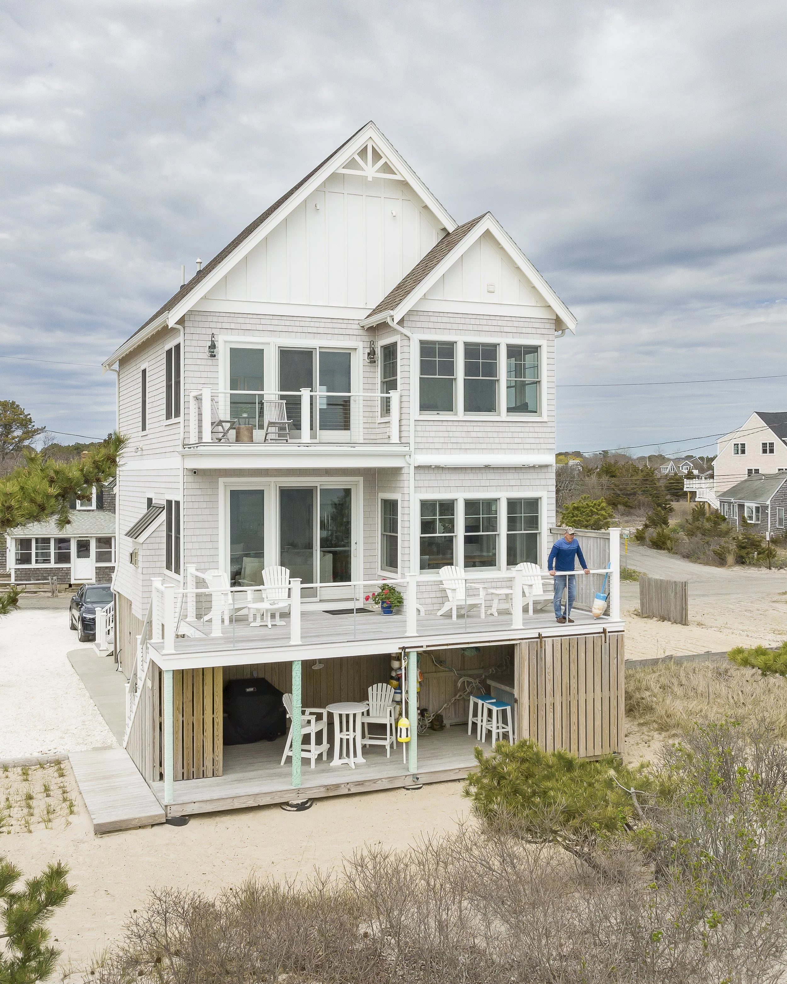 A multi-story white beach house with a wooden deck and patio furniture, overlooking a sandy area with sparse vegetation, under a cloudy sky.