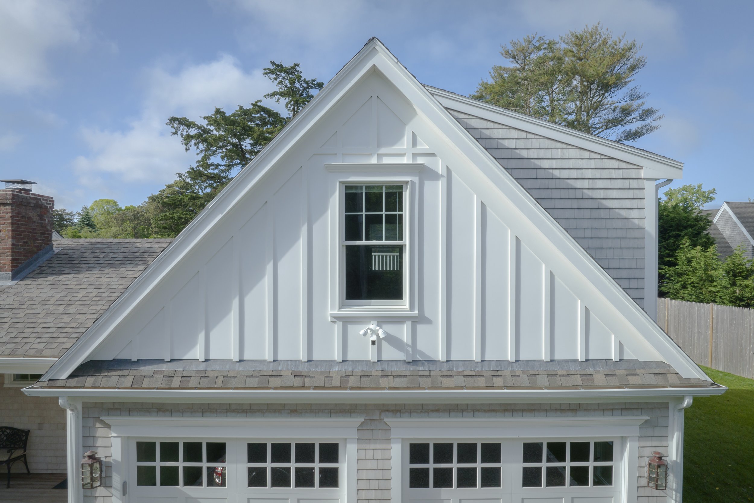 Front view of a modern house with a steep gable roof, white siding, a window, garage doors, and a chimney on the left side, with trees and a fence in the background.