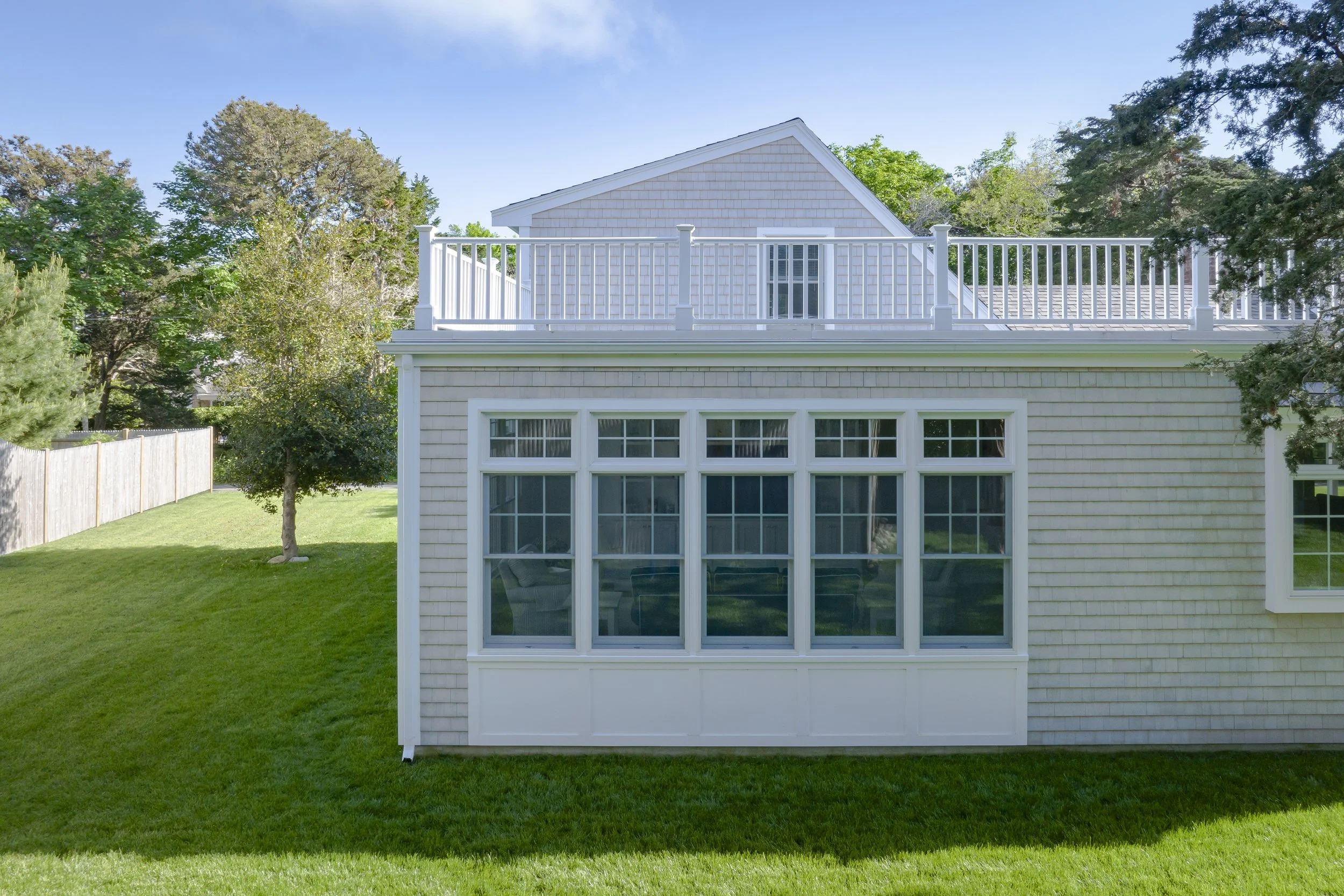 Back of a house with an enclosed porch, large windows, and a second-floor balcony with a white railing, surrounded by green grass and trees.
