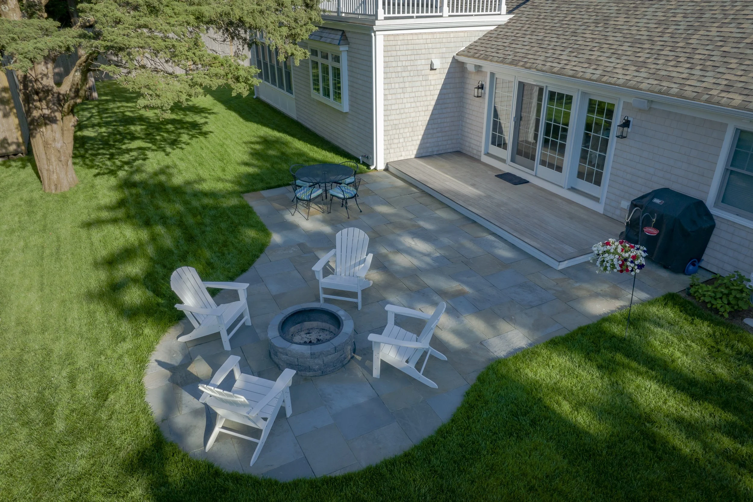 A backyard patio with four white Adirondack chairs arranged around a fire pit, a metal table with four chairs, a flower decoration, a barbecue grill, and a wooden deck attached to a house with sliding glass doors. The patio is paved with stone tiles,