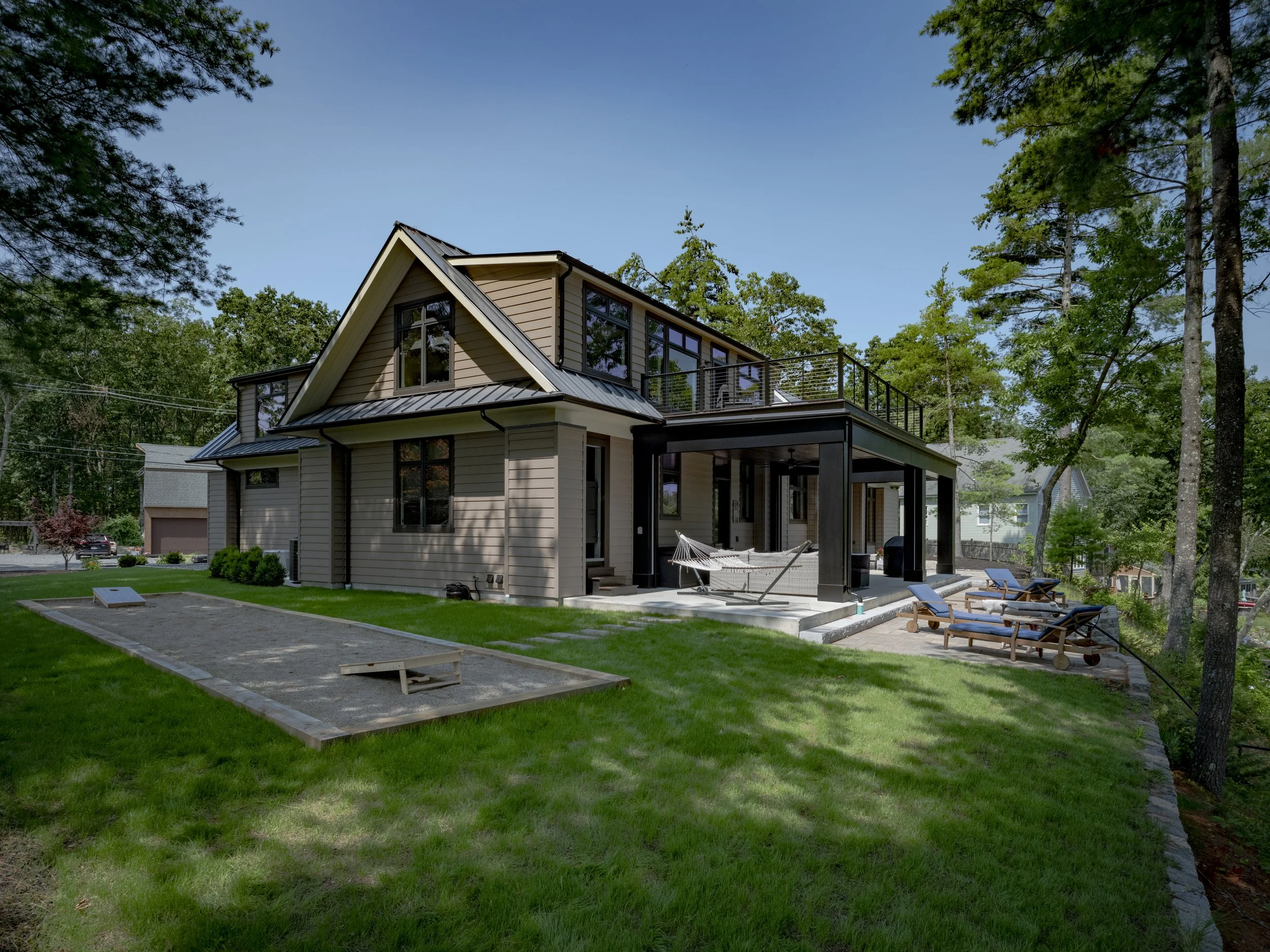 A modern two-story house with beige siding, black trim, and large windows, surrounded by green lawn and tall trees, with outdoor seating and lounge chairs on a patio.