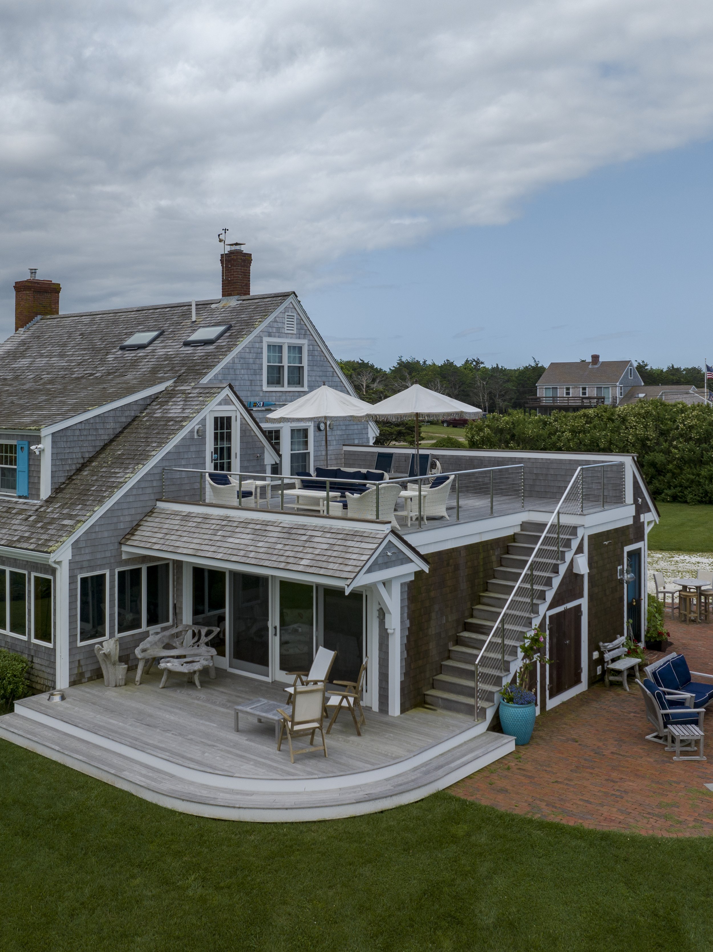 A two-story house with a rooftop deck featuring outdoor furniture and umbrellas, a wooden patio with chairs and tables, and a green lawn, under a partly cloudy sky.