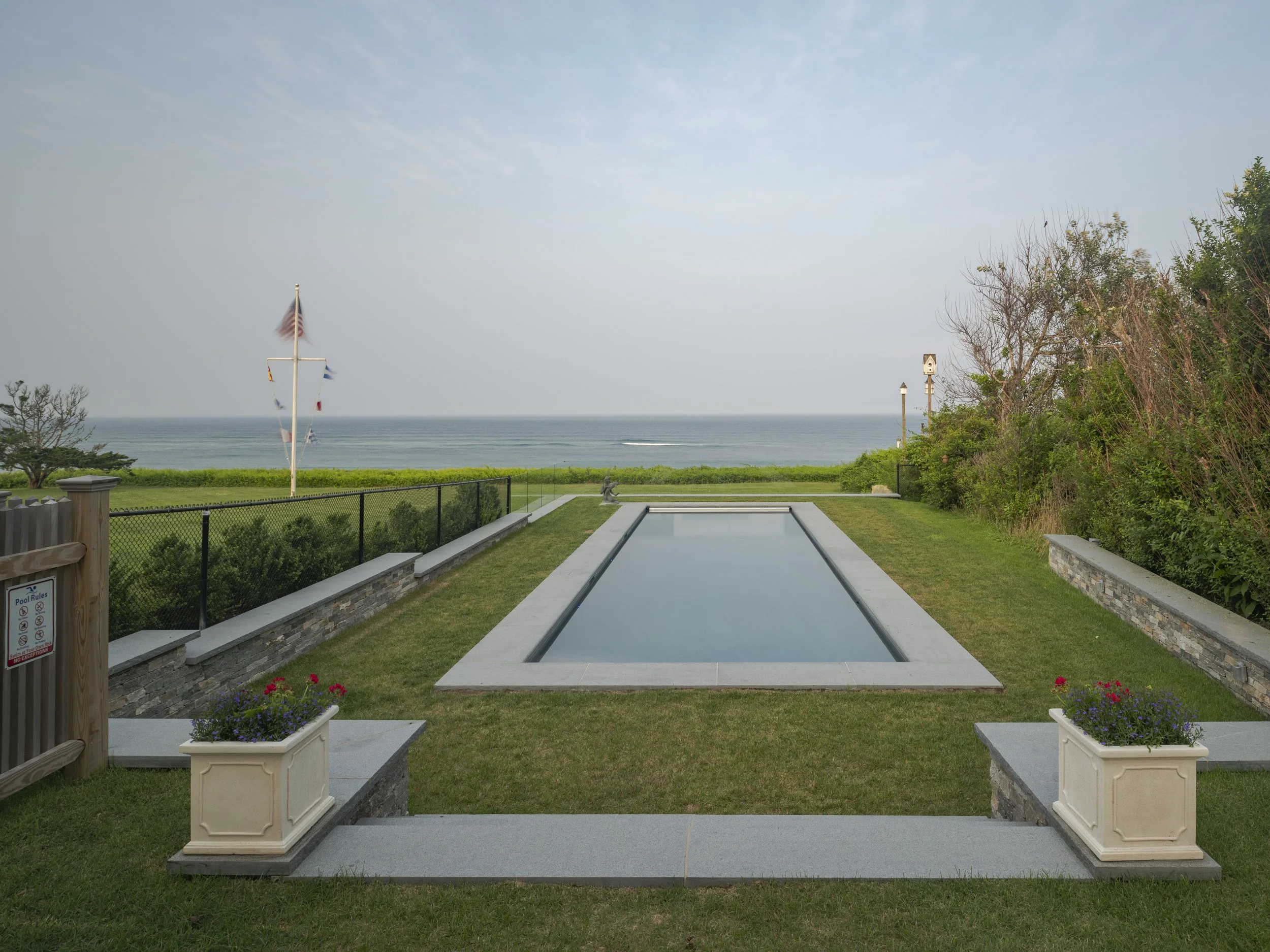 A rectangular swimming pool on a grassy yard, with the ocean and sky in the background. The yard is enclosed with a black fence, and there are potted flowers and trees.