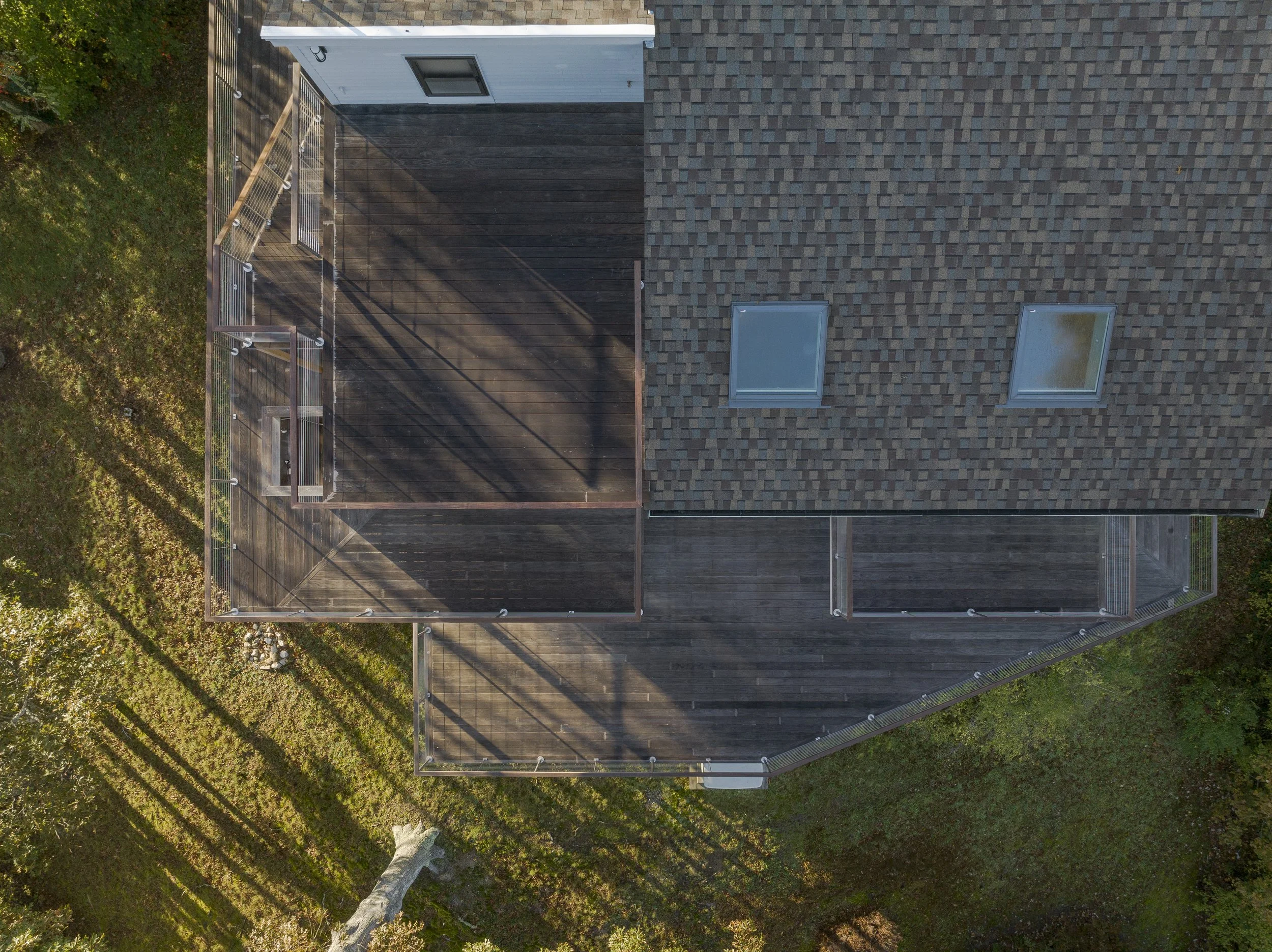 An aerial view of a house's roof and deck, with two skylights on the roof and a fenced balcony on the deck, surrounded by grass and trees.