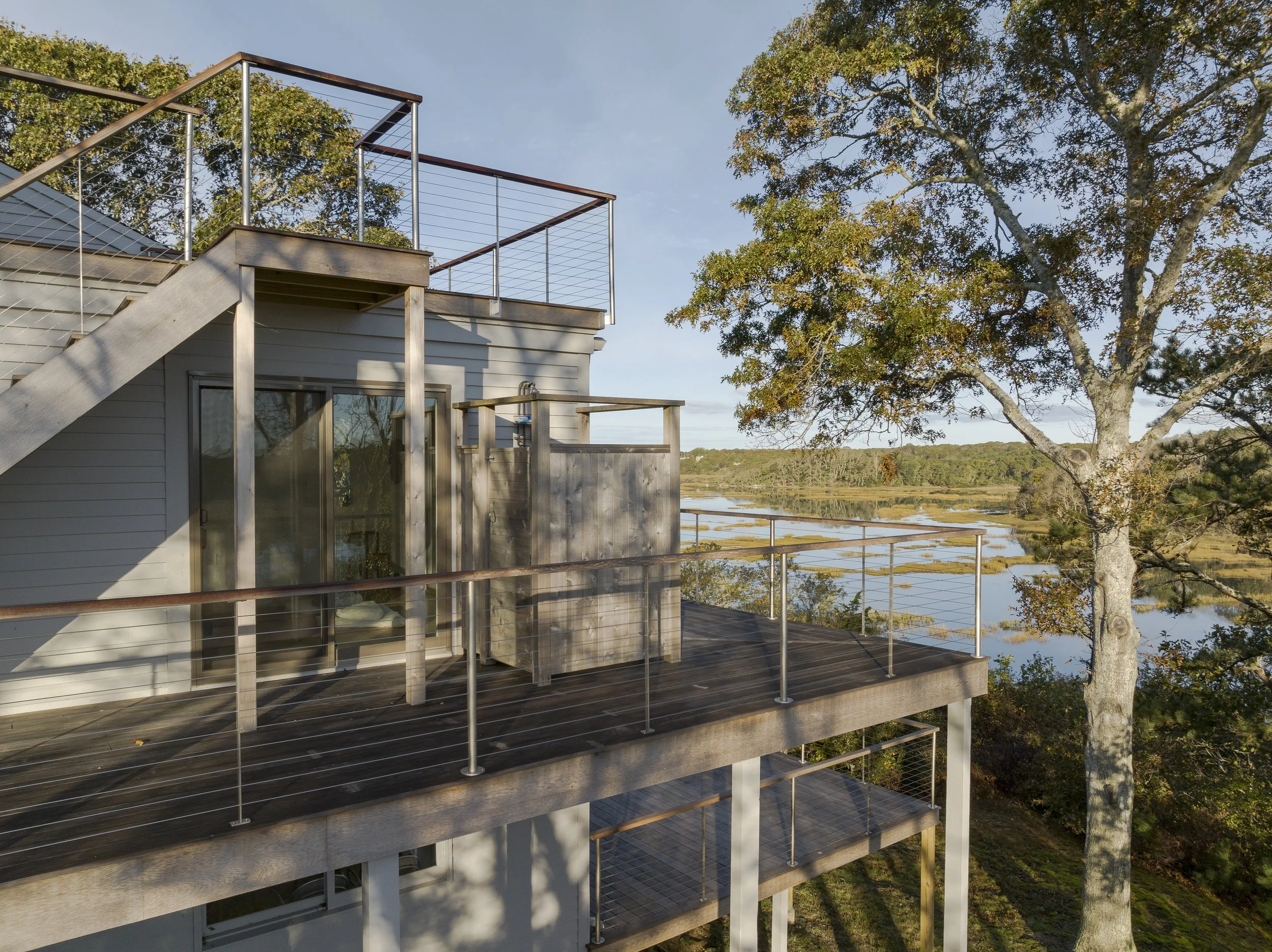 A multi-story house with a balcony, metal railings, and wooden decking surrounded by trees, overlooking a body of water and a marshy landscape with distant trees.